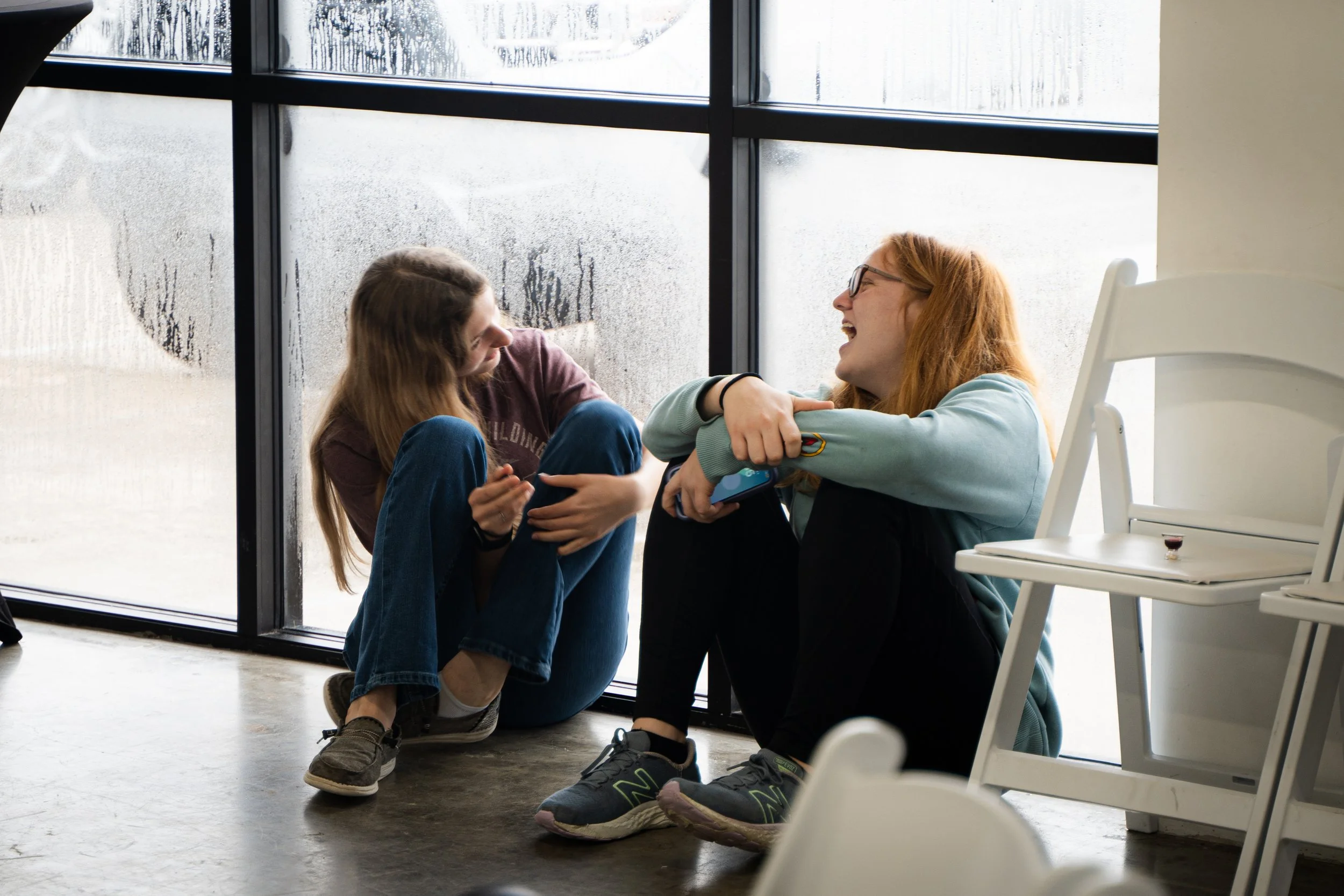 Two people sitting on the floor, laughing and talking, near a misty glass window.