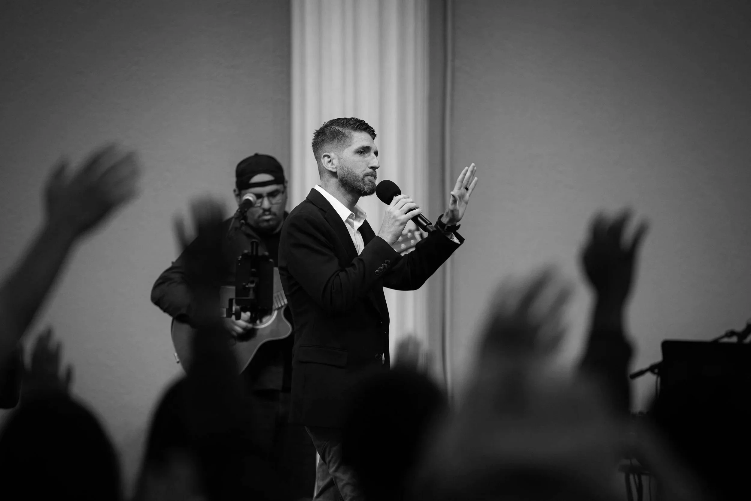 A man in a suit speaking into a microphone during a gathering, with another person playing guitar in the background. Audience members with raised hands are visible in the foreground.