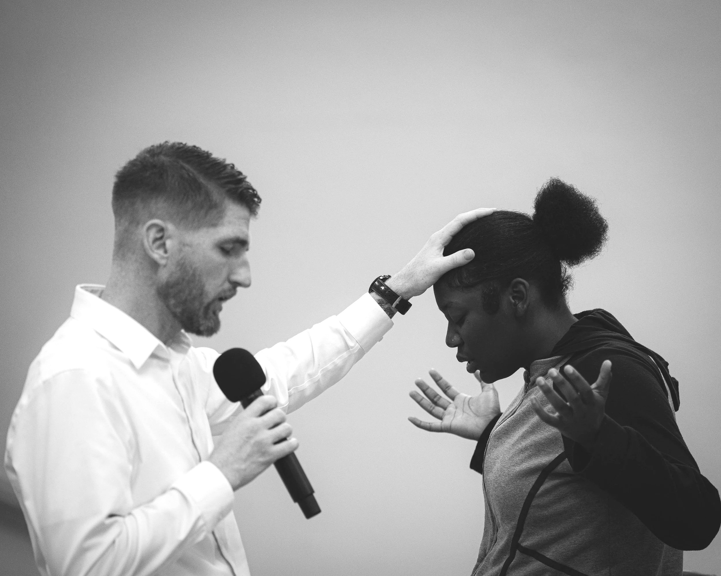 A man with a microphone places his hand on a woman's head in a gesture of prayer or blessing during a spiritual or emotional moment.