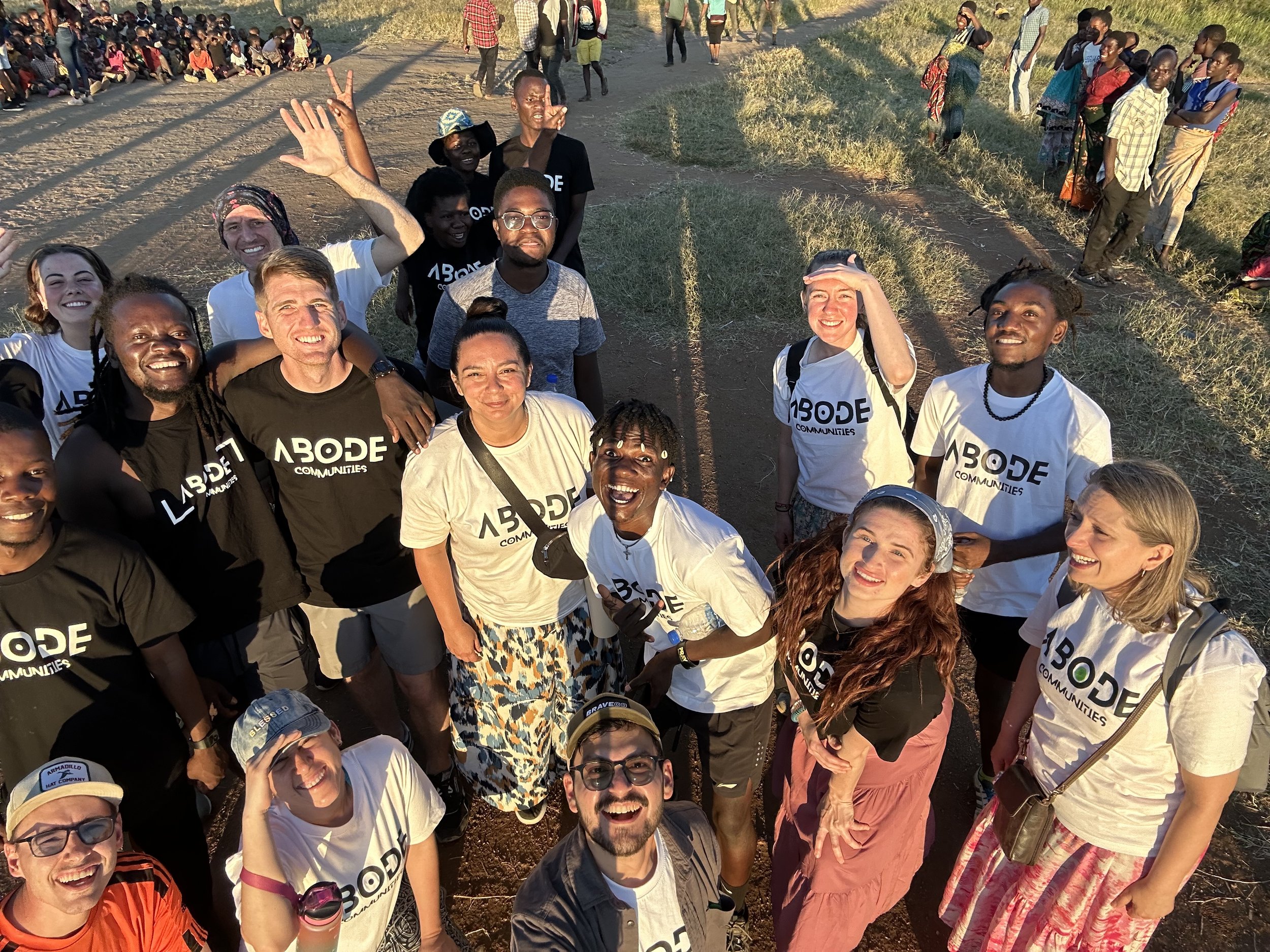 A group of people smiling and posing for a photo outdoors, wearing "ABODE Communities" t-shirts. The background shows a field with more people in the distance. The atmosphere appears warm and sunny.