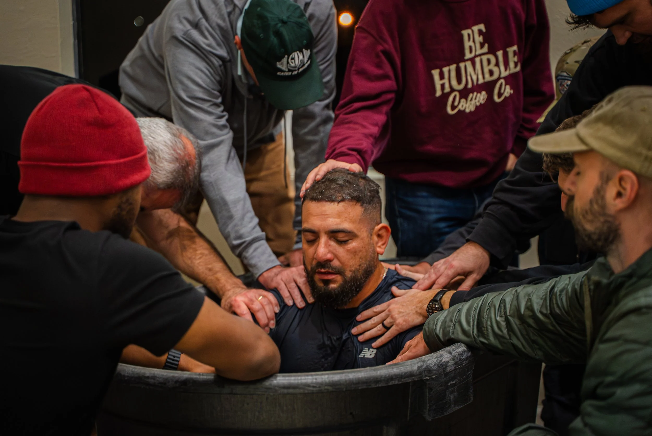 A man in a black shirt and a man in a red beanie are holding a man with short hair and a beard in distress during a baptism or religious ceremony, with several other people around supporting him.