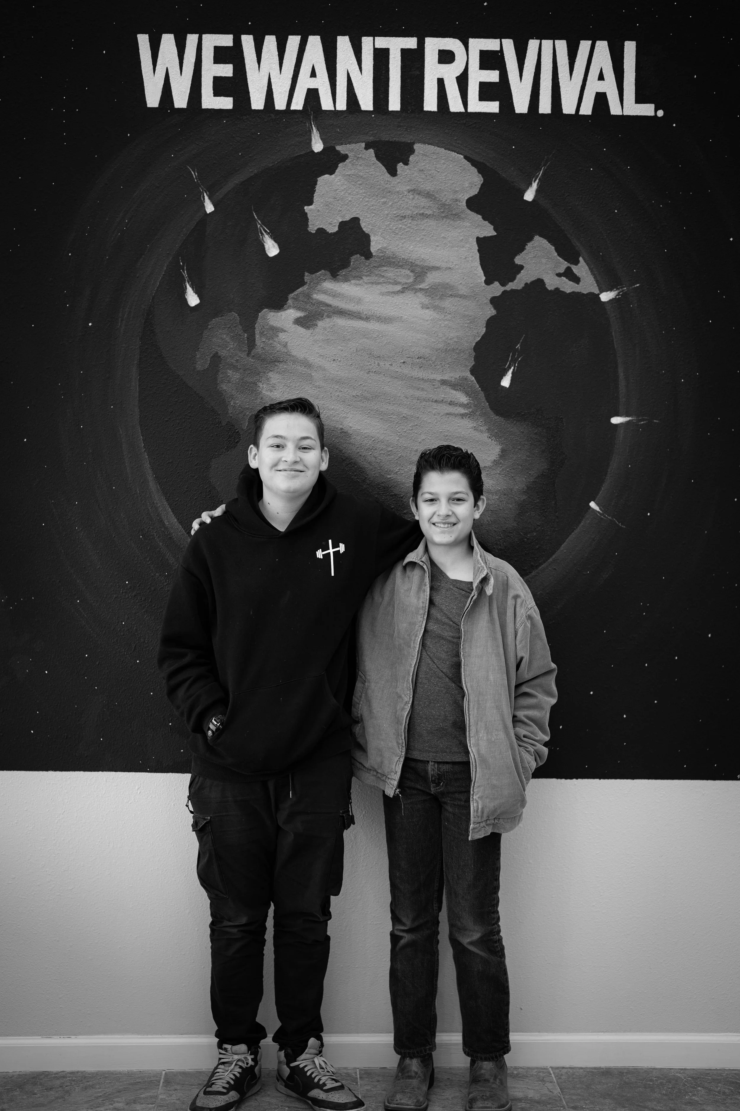 Two young boys standing in front of a mural with the words 'We Want Revival' and a depiction of Earth with meteorite impacts.