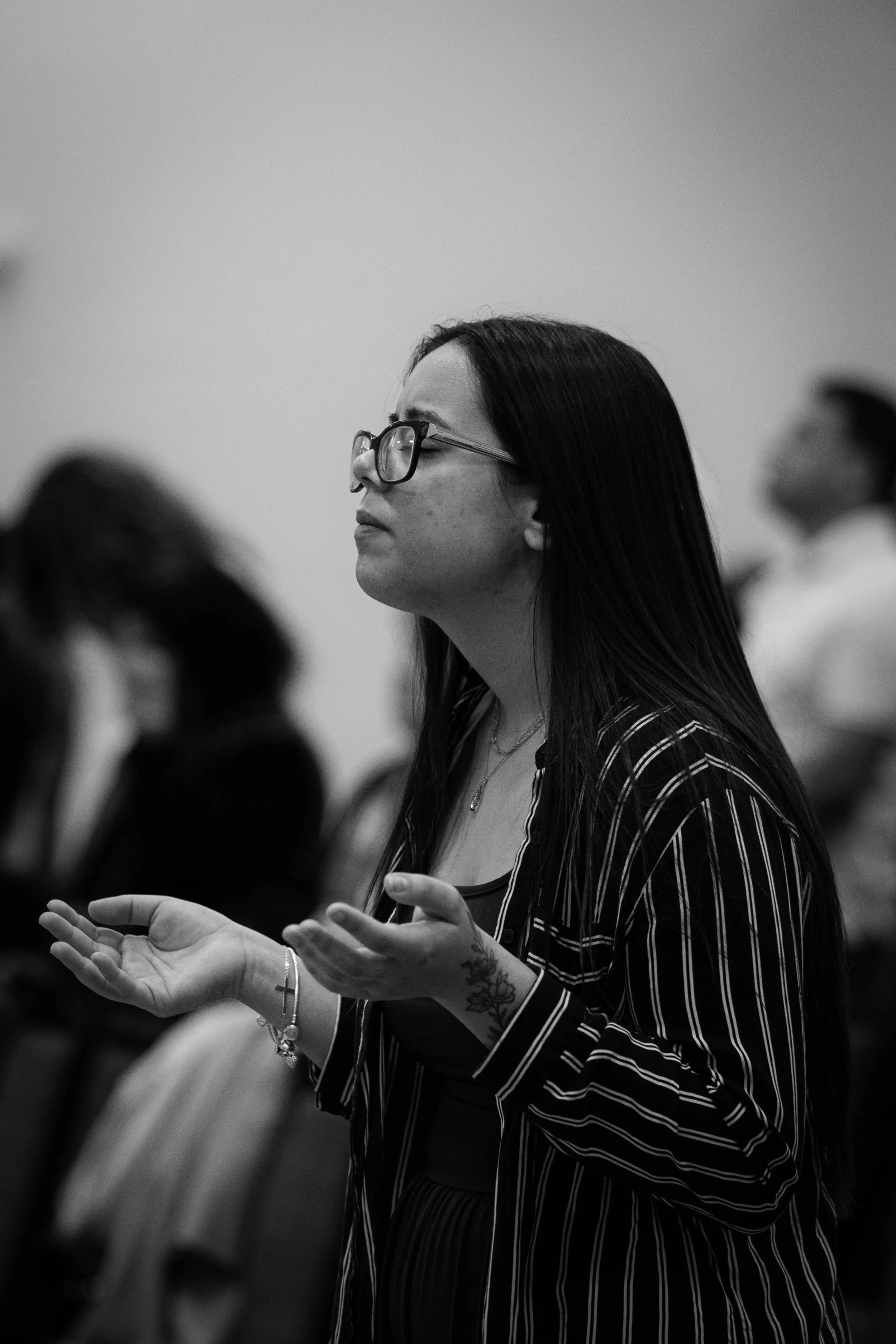 A woman with long dark hair, glasses, and tattoos on her wrist and arm, appears to be praying or meditating with her eyes closed and hands raised, in a room with other people.
