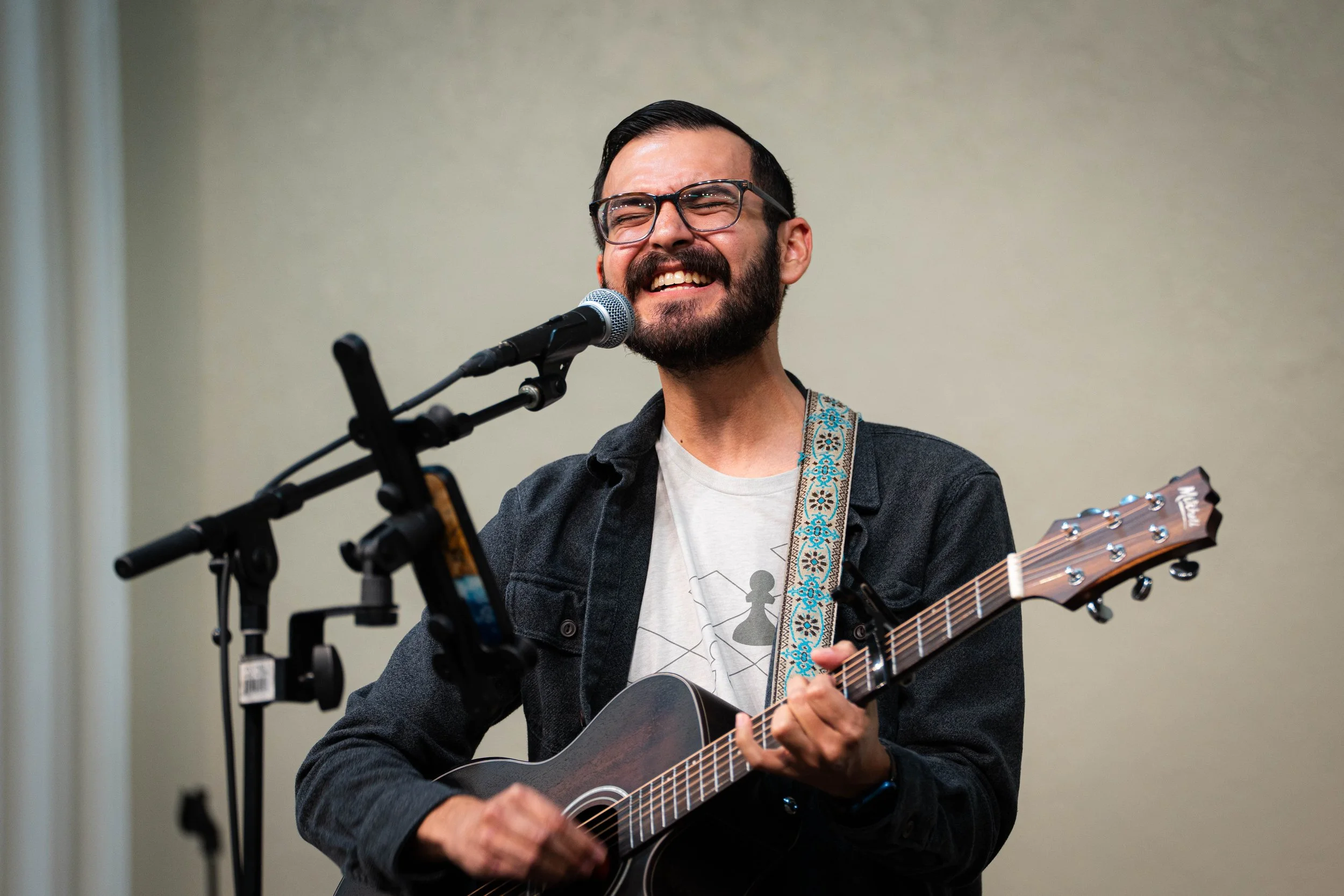 A man with glasses and a beard singing and playing an acoustic guitar, standing in front of a microphone.