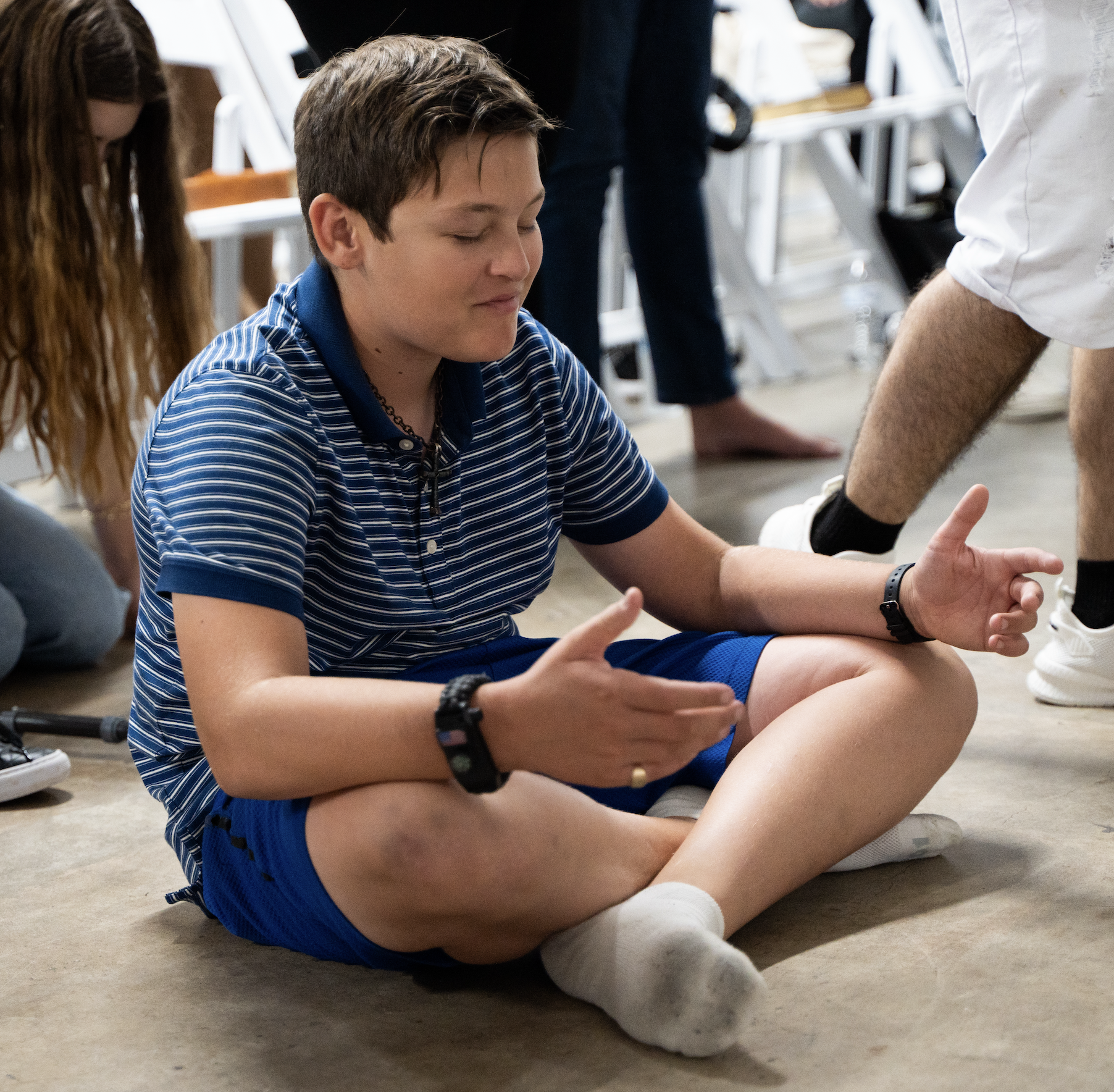 A young person sitting cross-legged on the floor with eyes closed, hands resting on knees in a meditative pose, wearing a blue striped shirt and shorts.