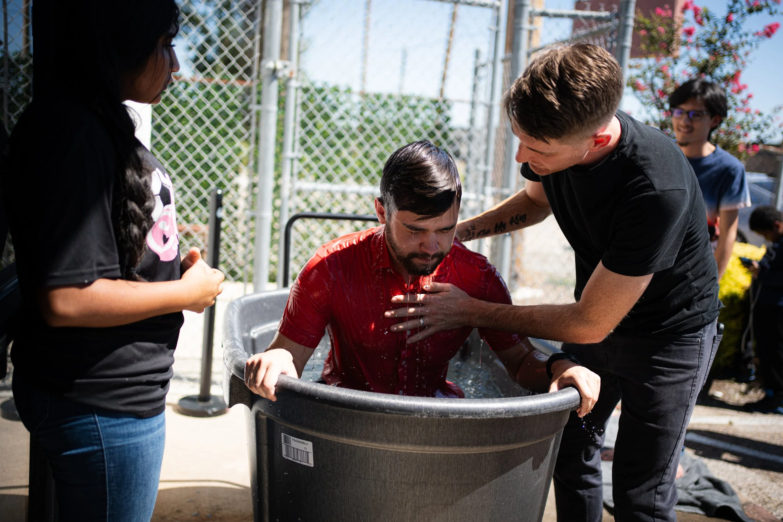 Man in a red shirt undergoing a baptism in a large metal tub, assisted by another person, with onlookers nearby.