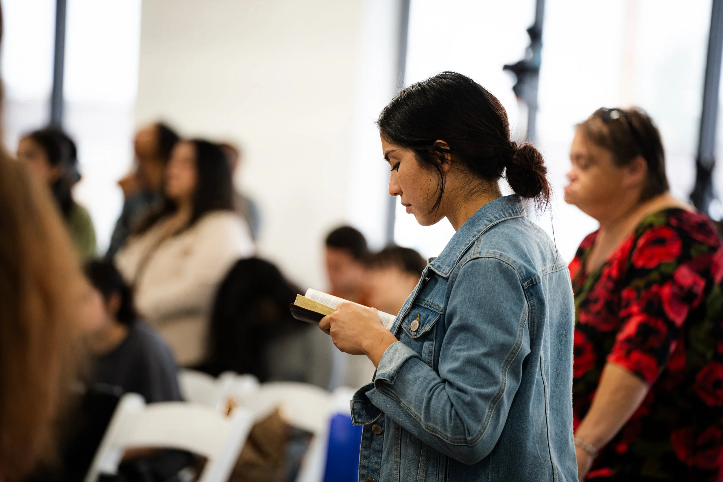 A woman in a denim jacket reading a book in a crowded room.