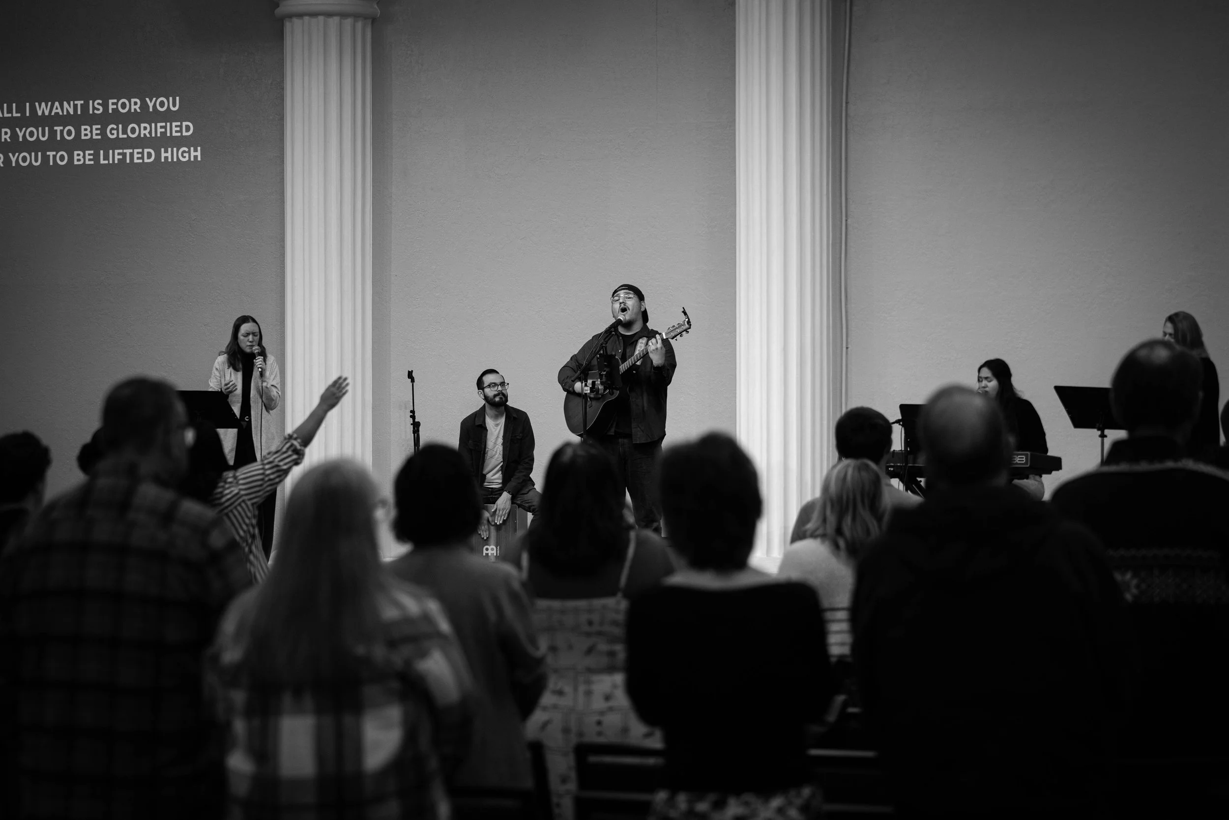 A worship band performing on stage with a man singing and playing guitar, surrounded by singers and musicians in front of an audience in a church or concert hall, in black and white.