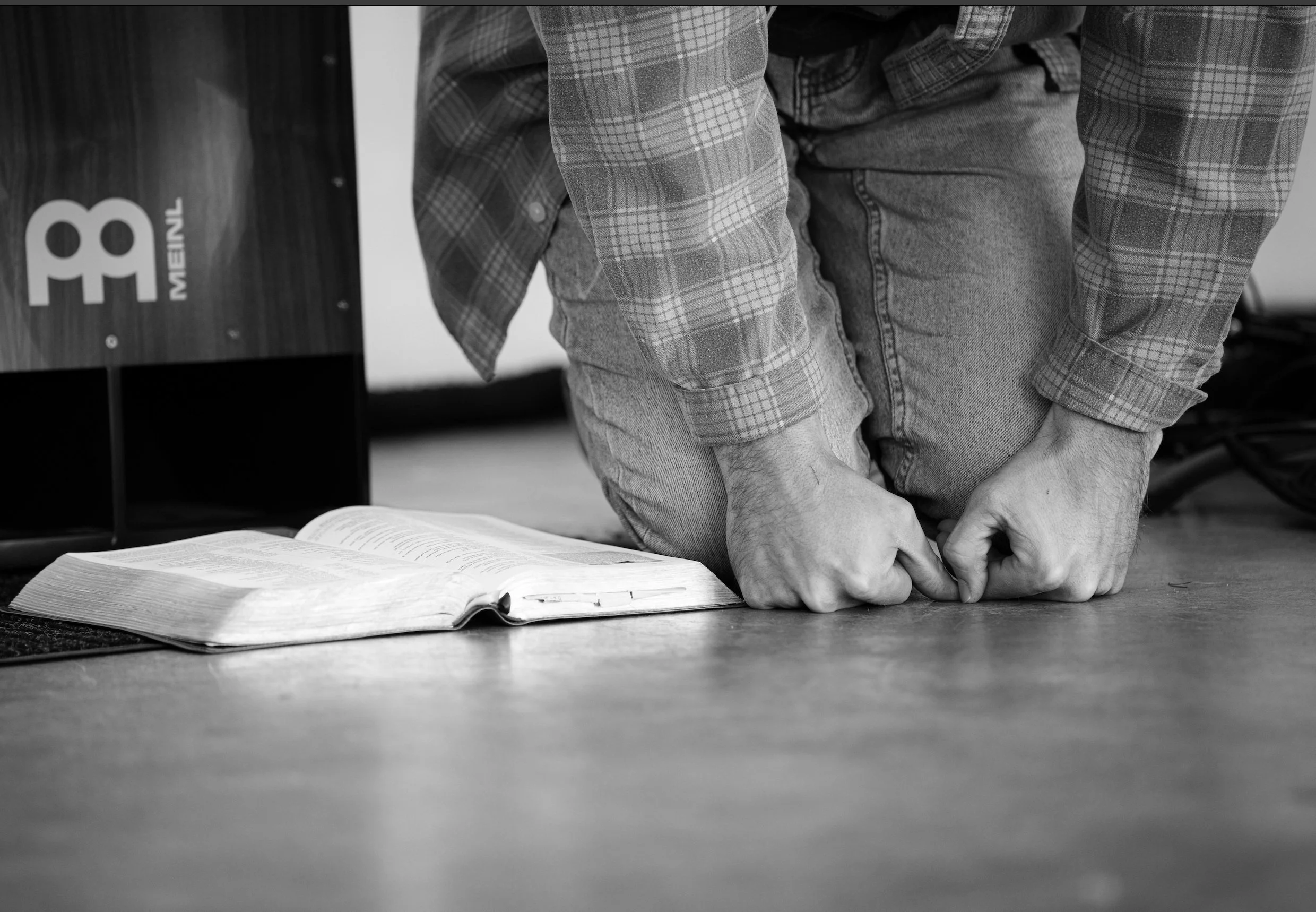 Person kneeling on the floor with open Bible in front, fists clenched and head bowed, in a black and white photo.