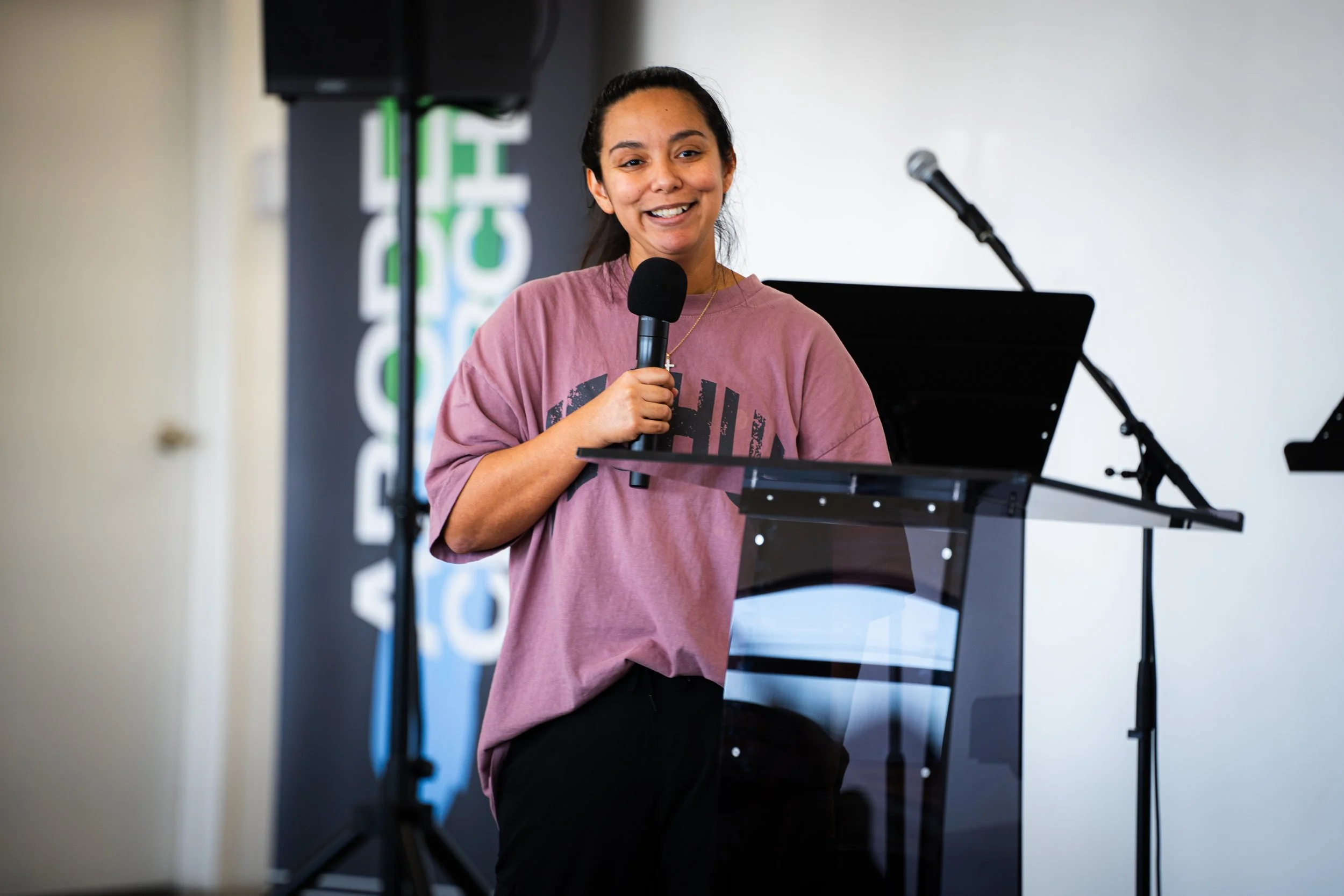 Young woman with dark hair in a ponytail speaking into a microphone at a podium, in front of a logo and a microphone stand.