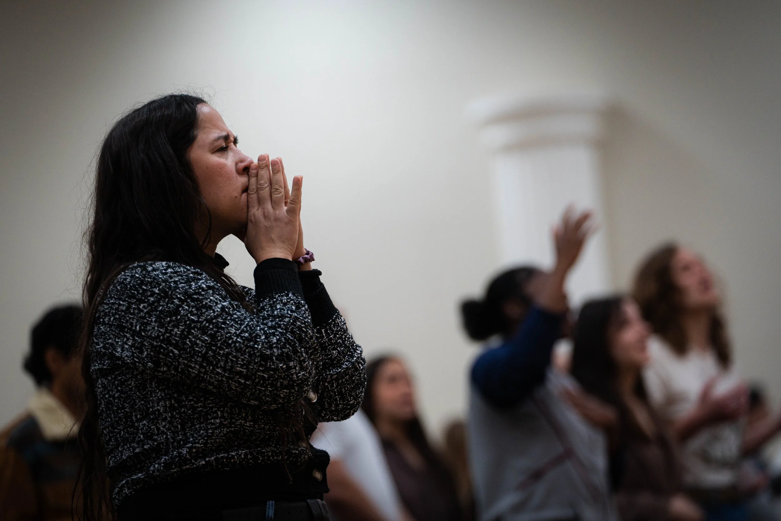 A woman with long dark hair is praying or expressing emotion with her hands covering her mouth, in a room with other people who are seated, some raising their hands.