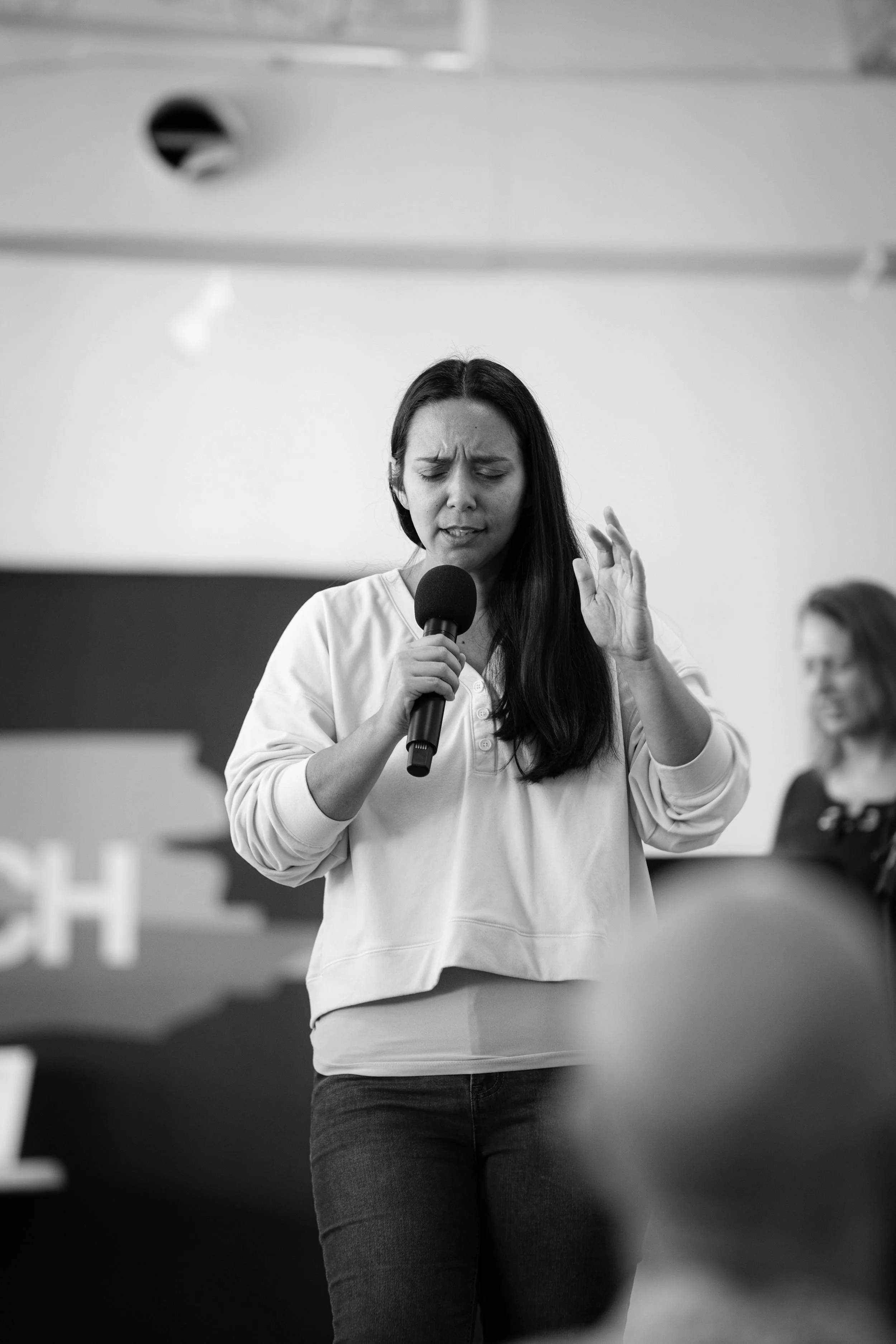 Black and white photo of a woman speaking into a microphone while standing, with eyes closed and one hand raised, in an indoor setting with another person blurred in the background.