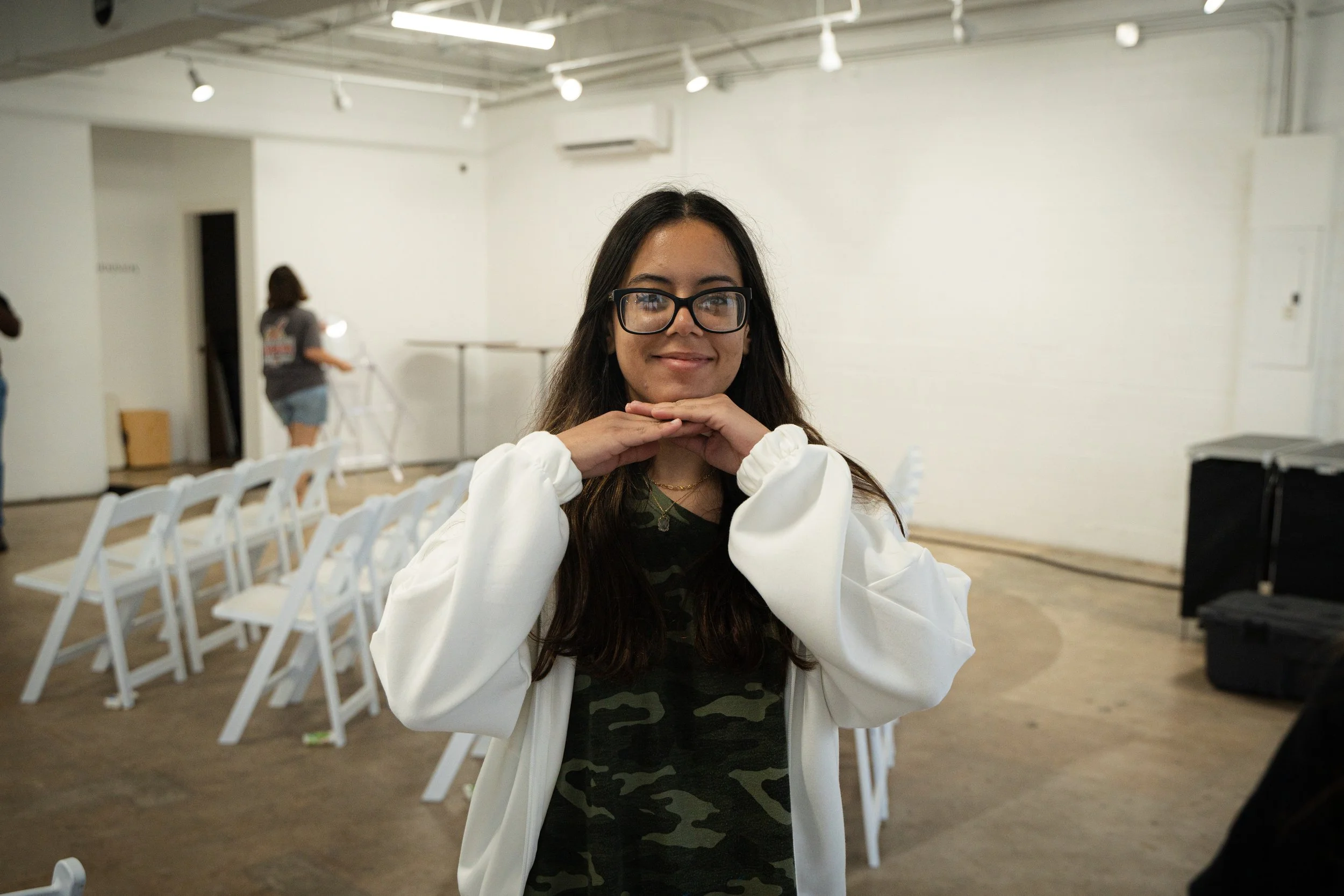 Young woman with glasses smiling, wearing a white jacket and camouflage shirt, posing with her hands under her chin indoors, with chairs and people in the background.