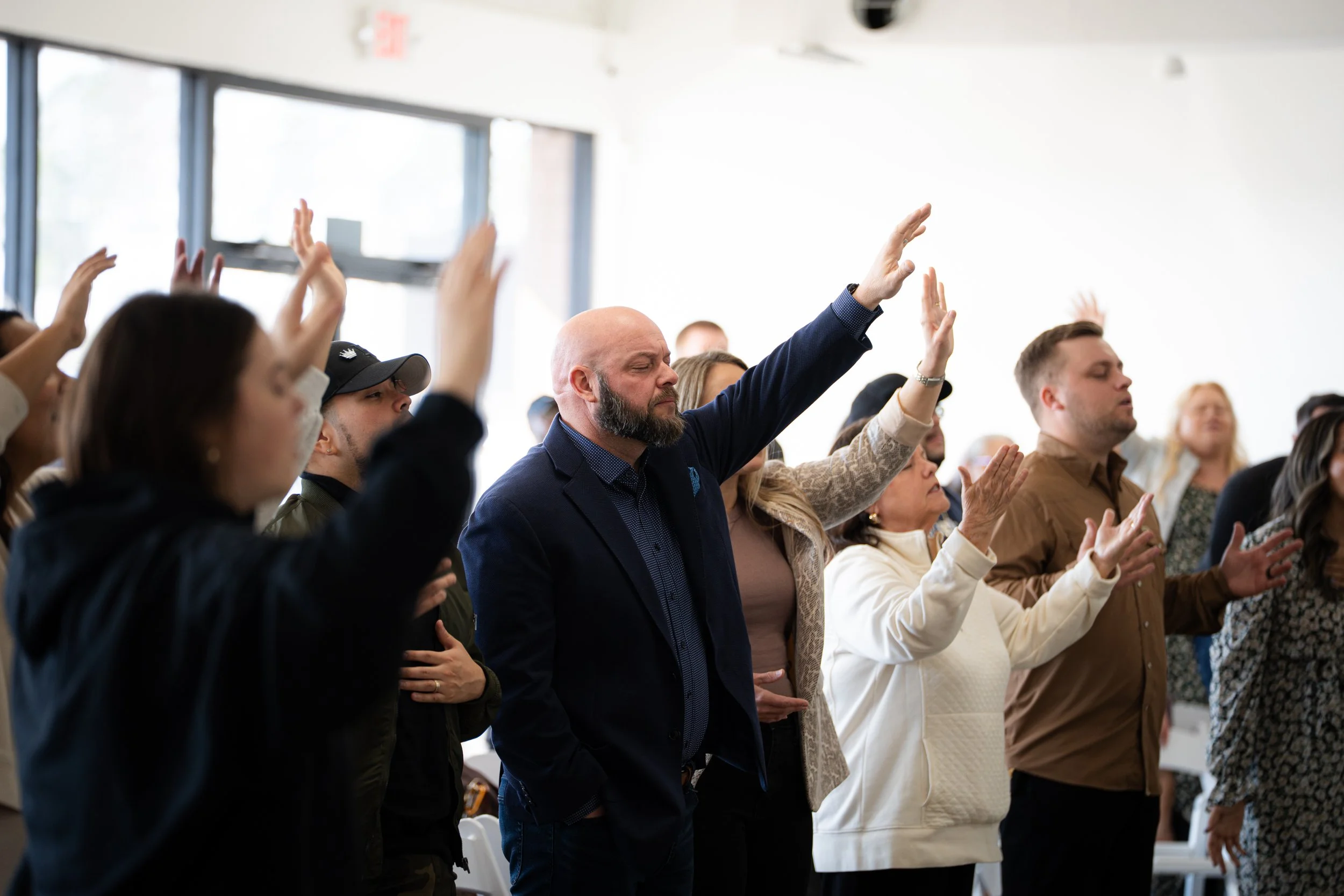 People standing in a room with bright natural light, some with eyes closed and hands raised, appearing to be in prayer or worship.