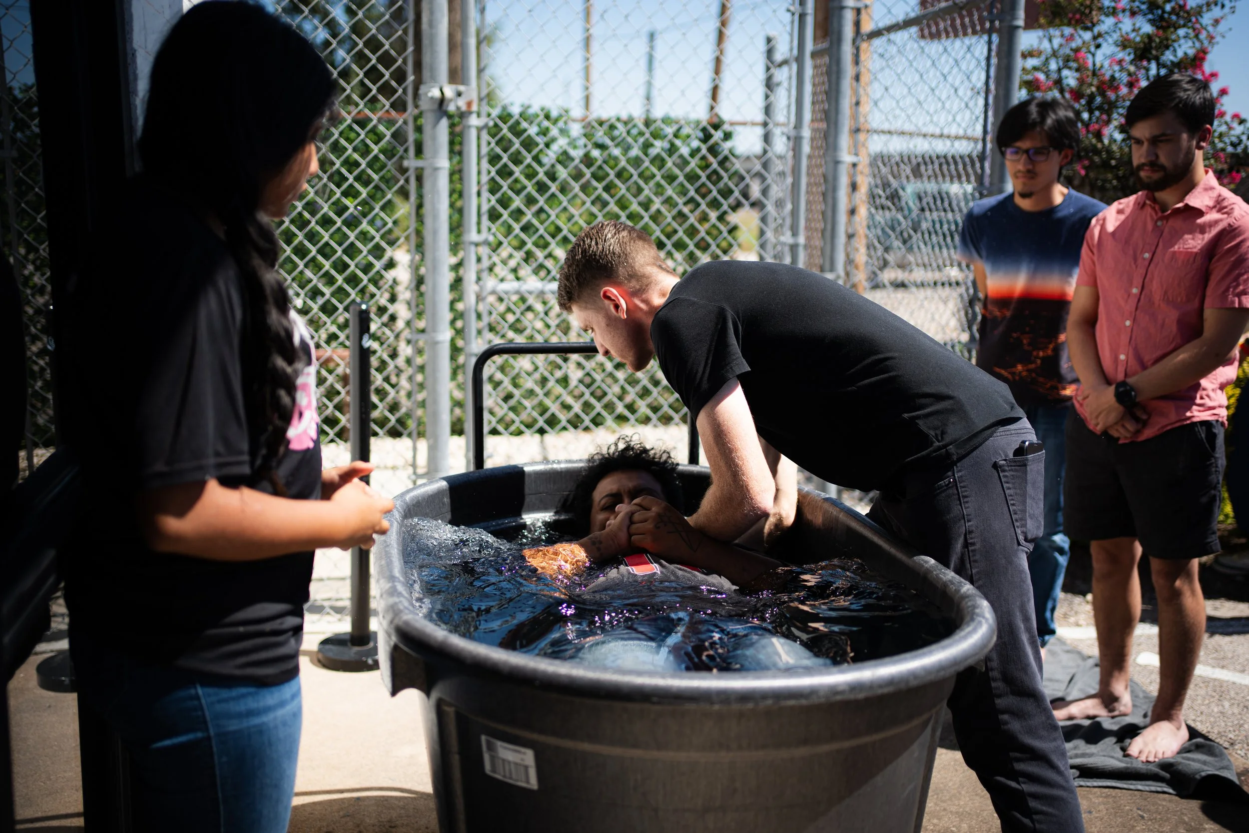 Person being baptized in a large tub outdoors, surrounded by onlookers.