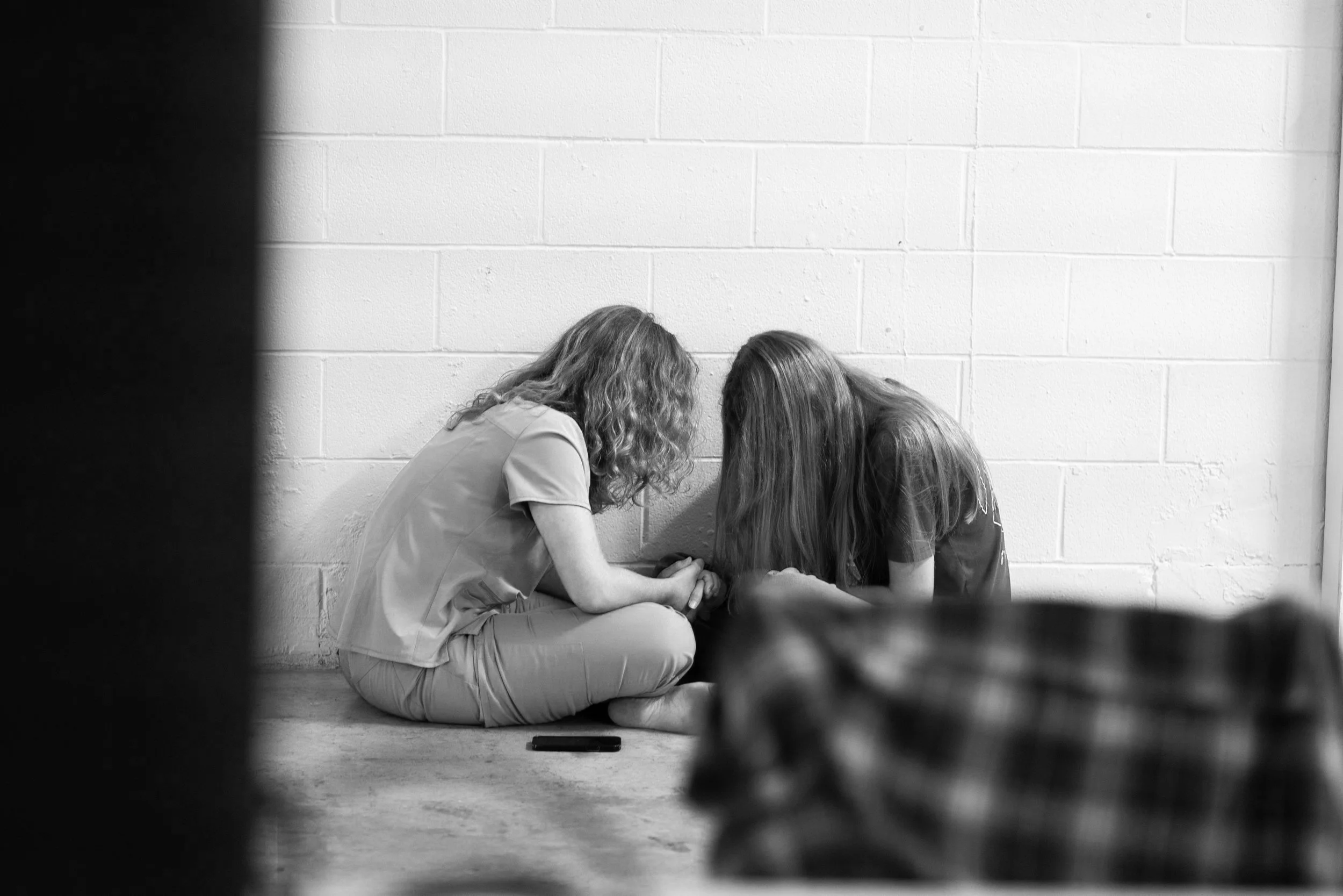 Two individuals sitting on the floor in front of a white brick wall, heads bowed and hands clasped together in a gesture of support or prayer.