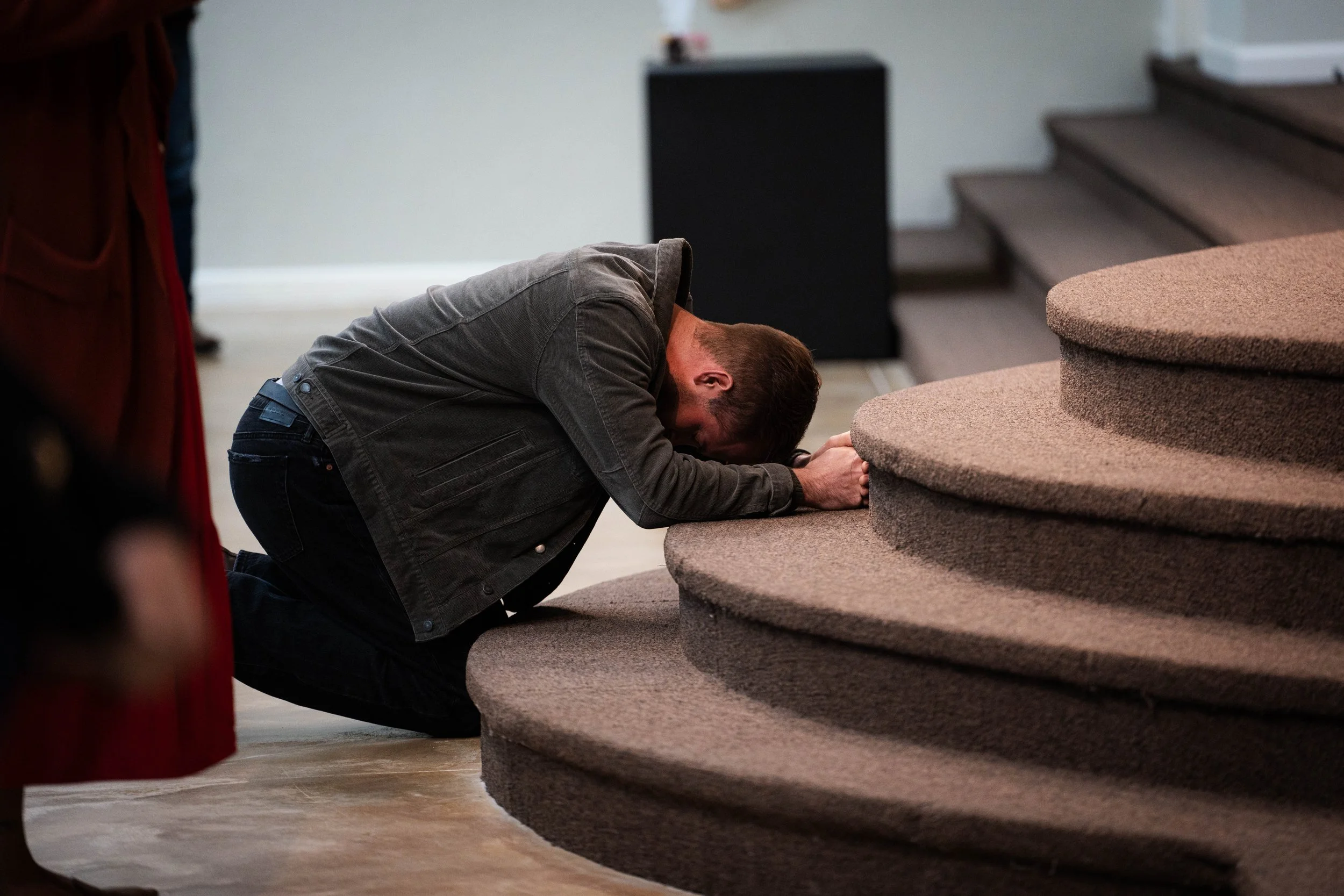 A man kneels on the floor, bowing in prayer with his forehead touching the carpeted steps of a staircase.