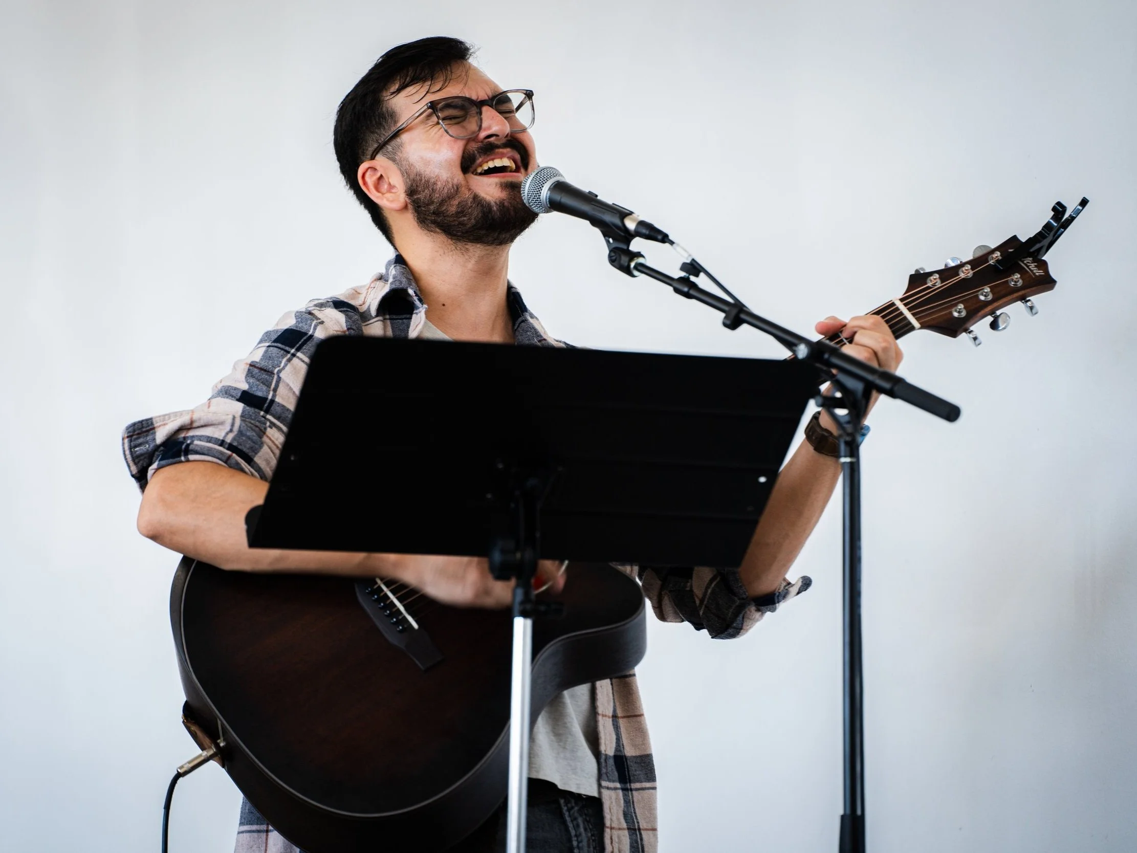 Man with glasses singing and playing acoustic guitar at microphone stand, wearing plaid shirt.