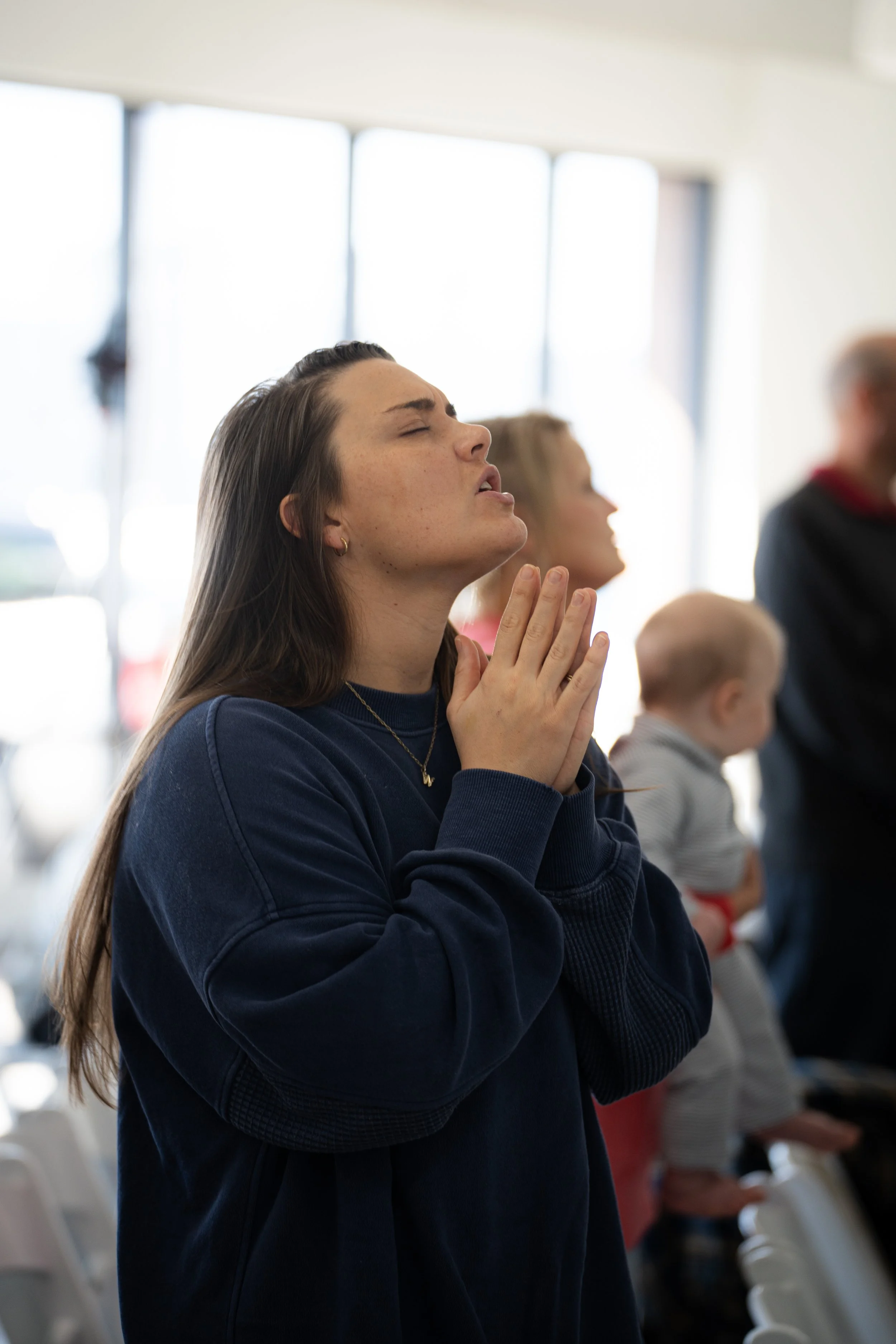 A person with long brown hair in a blue sweatshirt is standing with eyes closed and hands clasped in a prayerful gesture, surrounded by others in a softly lit room.