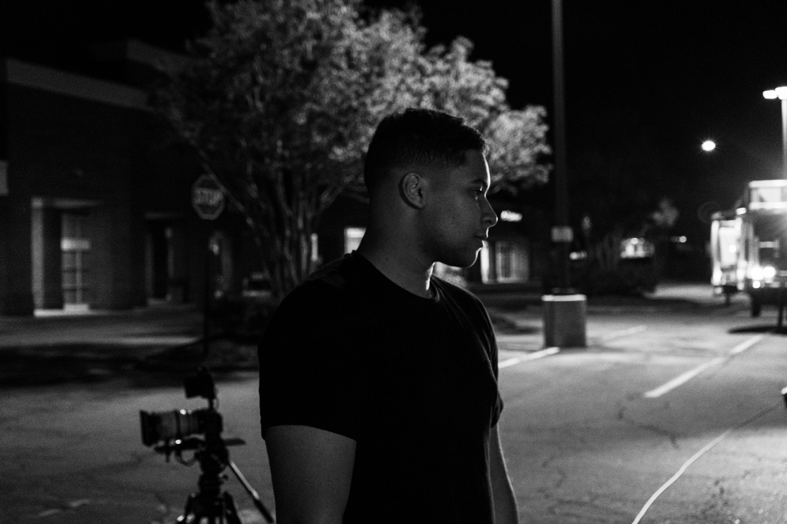 A young freelancer stands in a parking lot at night, facing right, with video equipment and some buildings in the background, illuminated by streetlights.