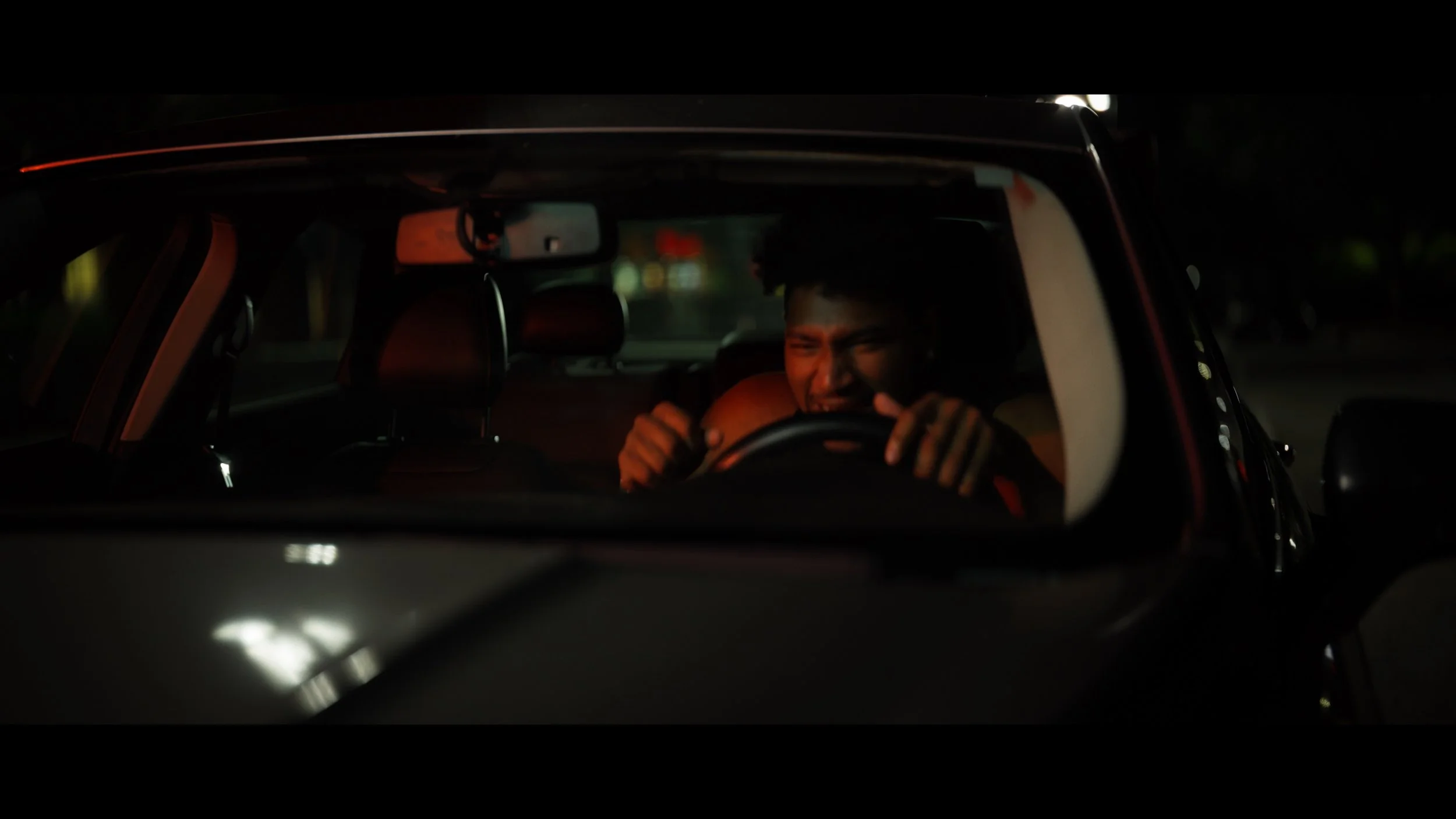 A person inside a car at night, gripping the steering wheel tightly and making a grimacing expression.