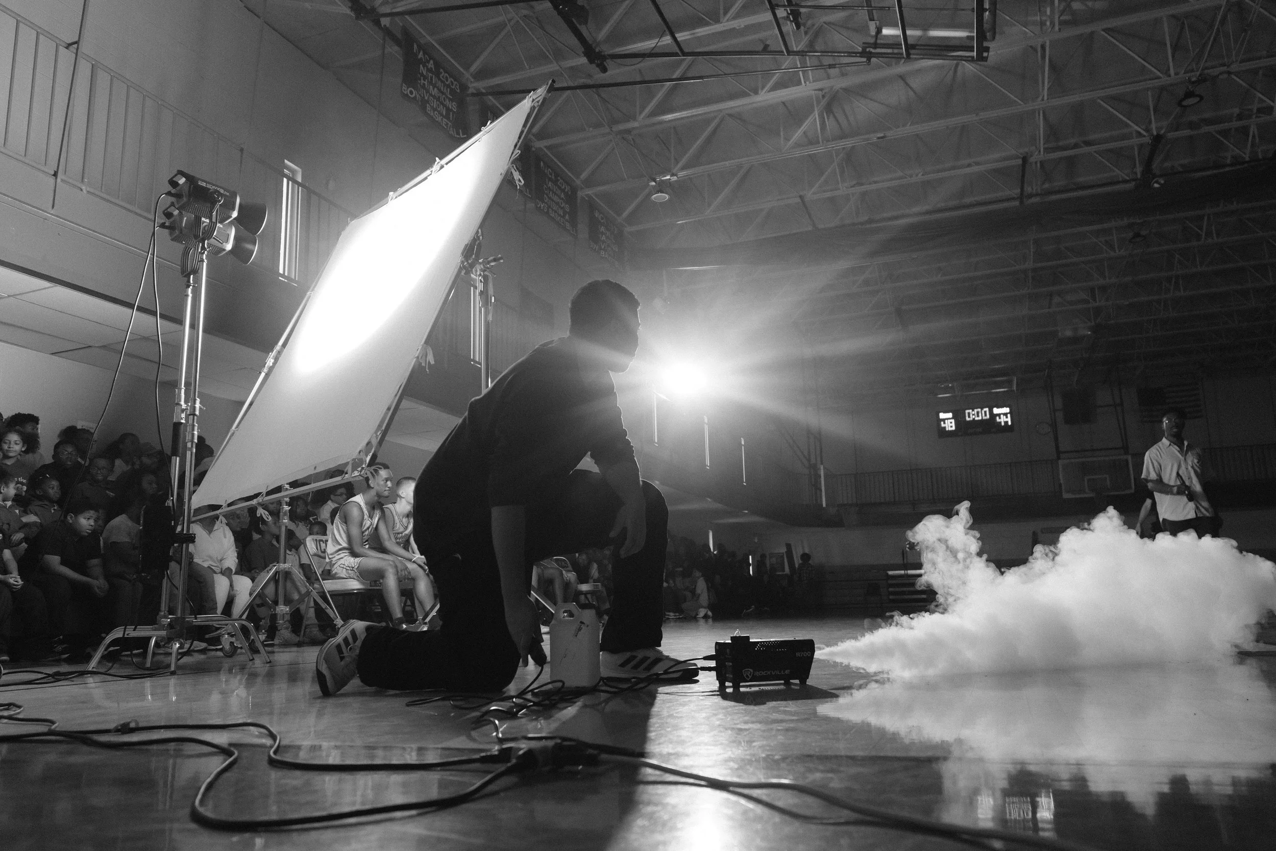 Kneeling video freelancer operating fog and video equipment on gymnasium floor for a commercial, with actors and extras sitting on the bleachers with scoreboard in background.