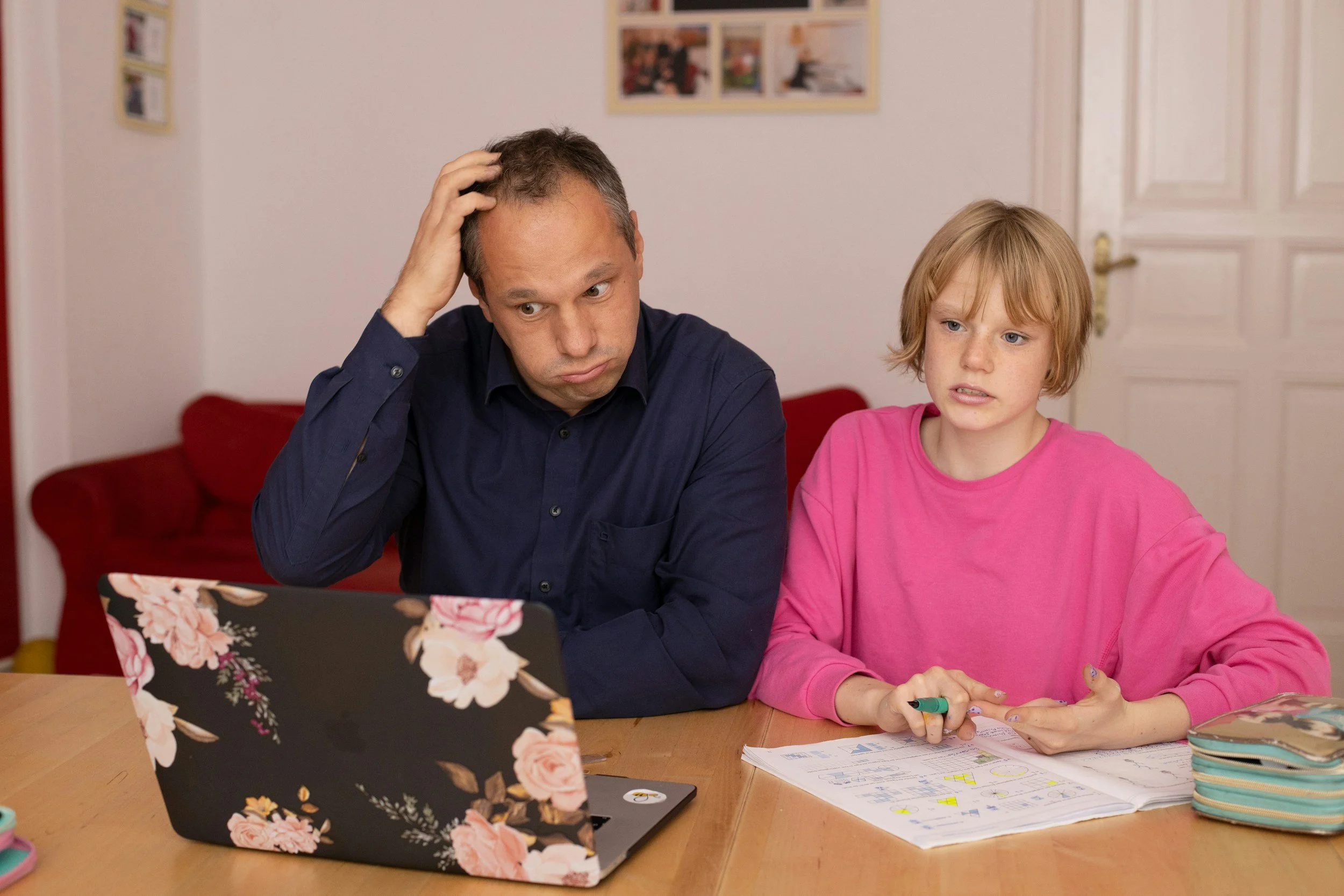 A man and a young girl sit at a table looking at a laptop and schoolwork with puzzled expressions.