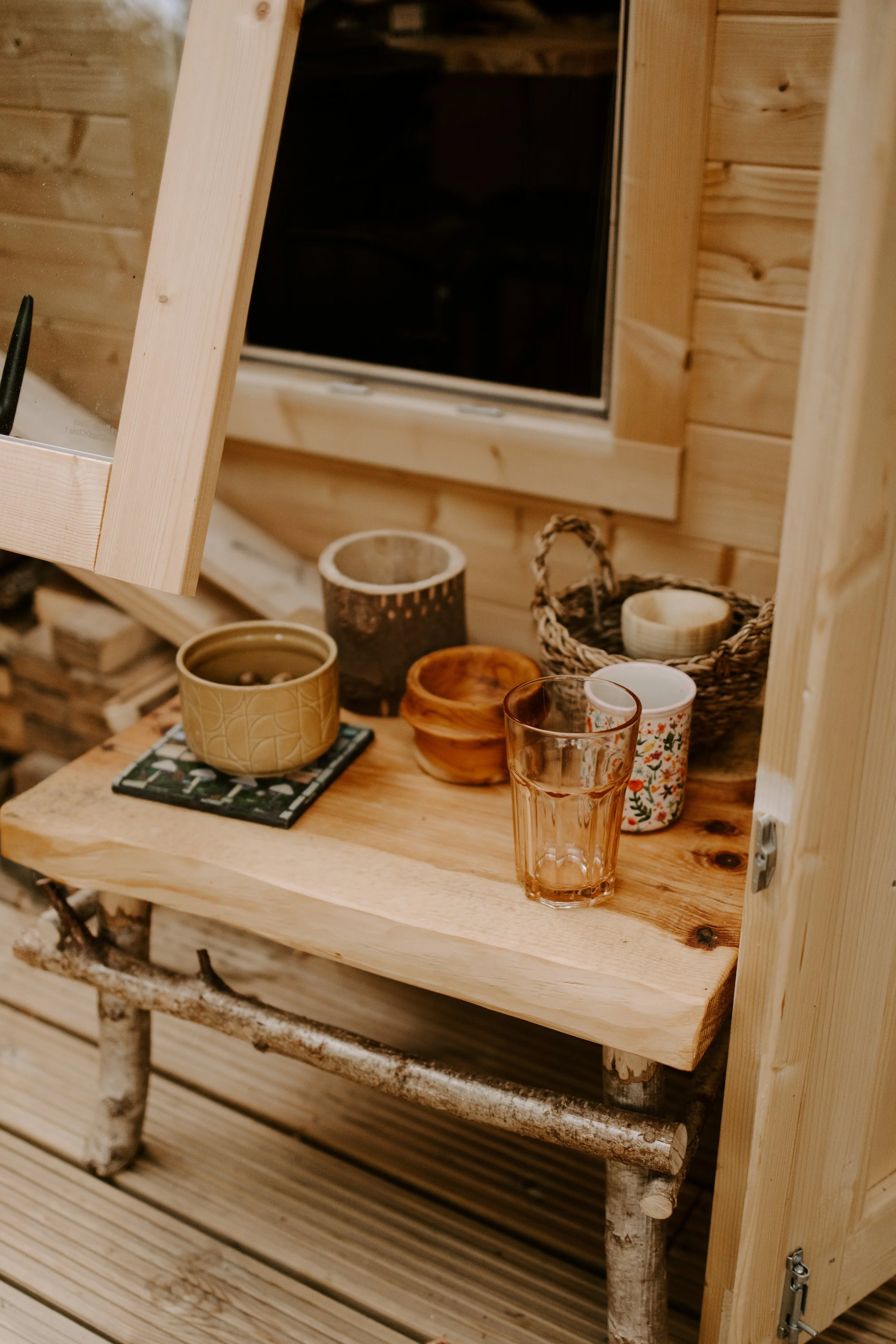 A wooden shelf with various cups and bowls placed near a window in a cozy, wood-paneled interior.