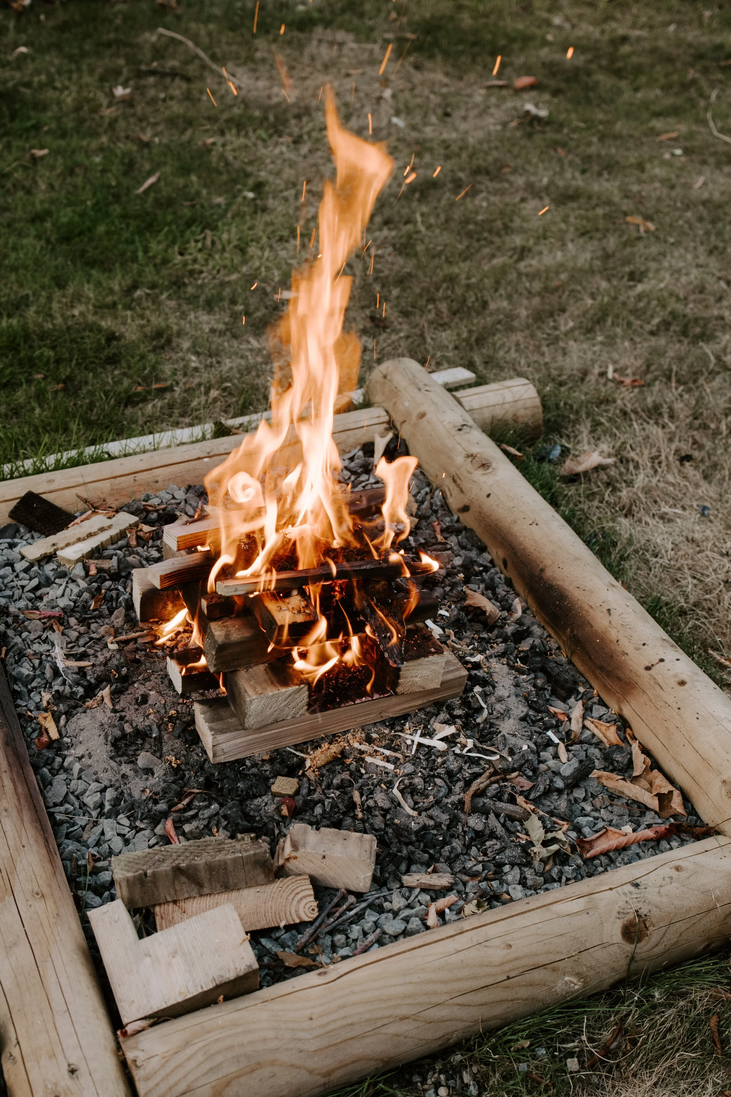 Fire burning in a small outdoor fire pit, surrounded by gravel and a wooden border, with some logs and grass around.