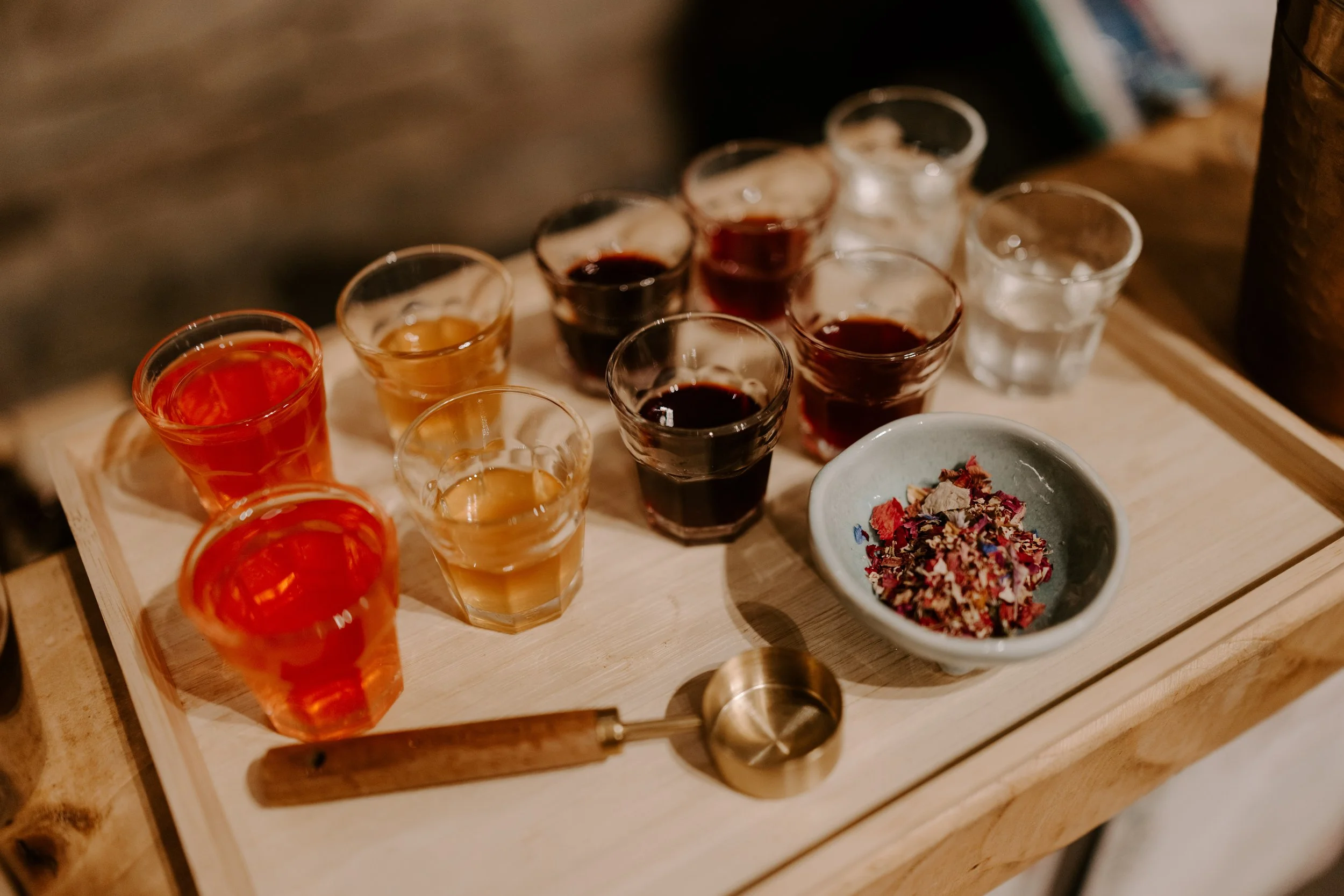 A wooden tray holding nine small glasses filled with various colored liquids, which are homemade foraged syrups and cocktail ingredients and a small bowl containing dried flower petals, with a measuring scoop nearby.