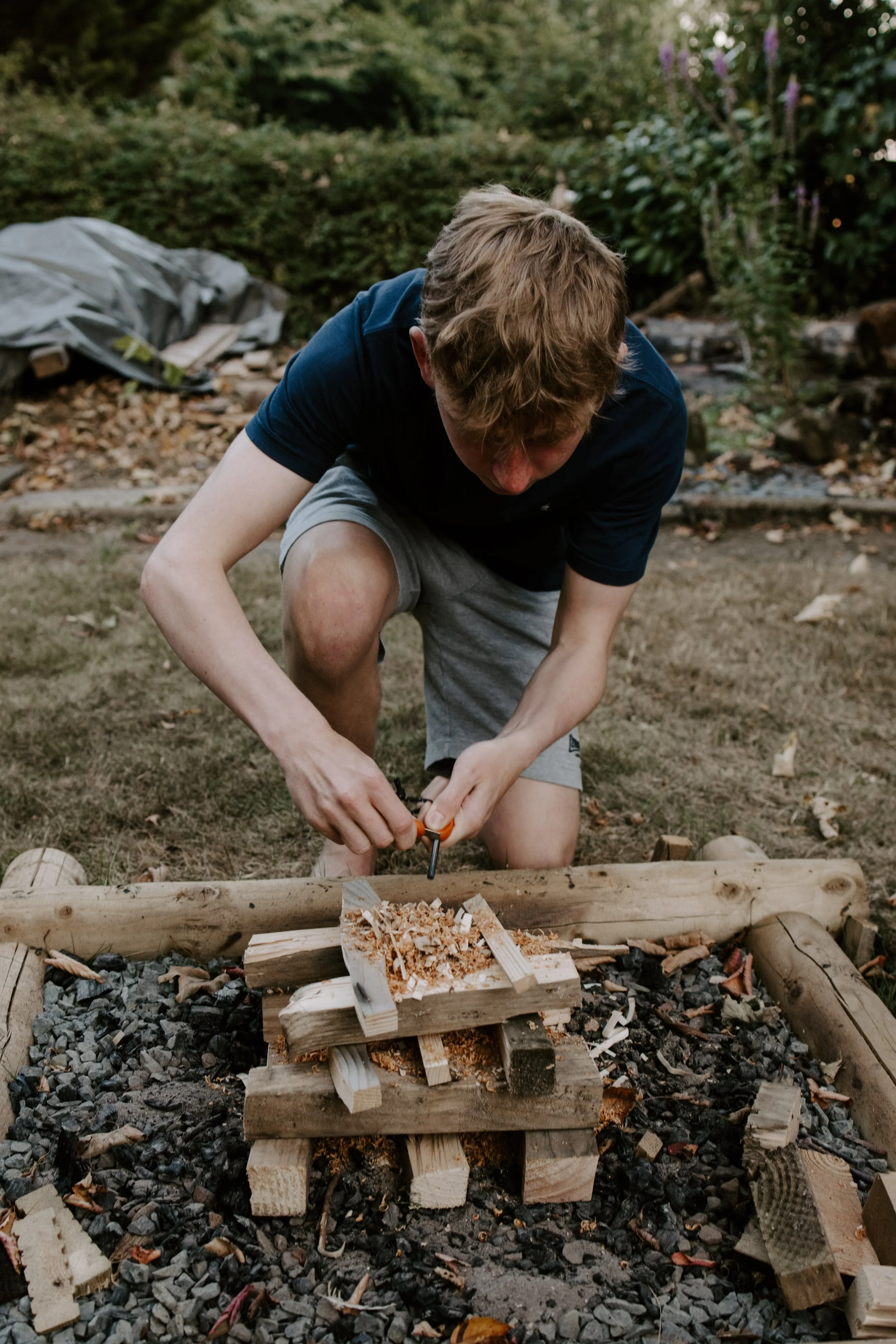 A teenage boy kneeling down by a campfire at Feral Outdoor Company space starting to use a ferro rod to start a fire.