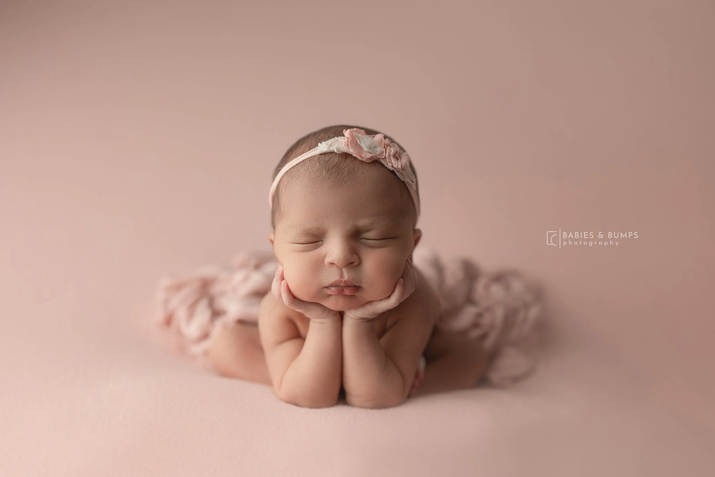 A newborn baby girl with closed eyes, wearing a pink headband with a bow, resting her chin on her hands on a pink background.