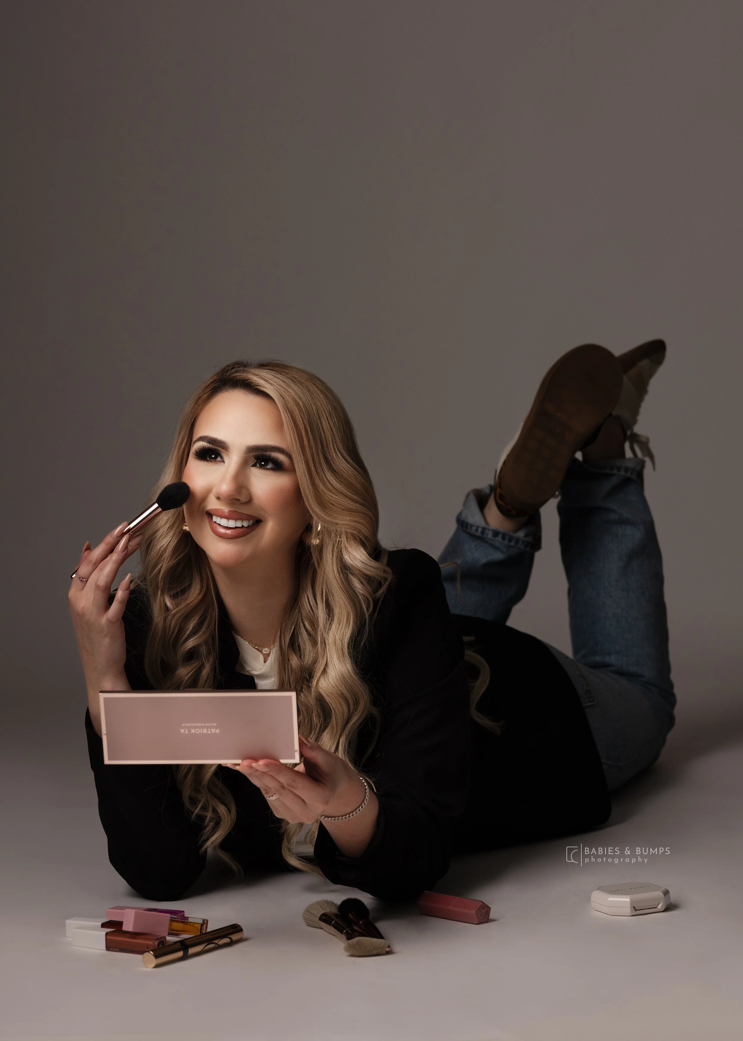 A woman lying on her stomach, smiling, holding a makeup palette while applying makeup. Her other hand holds a makeup brush. She has long wavy blonde hair and is wearing a black blazer. On the table in front of her, there are makeup products, brushes, and a pink palette.