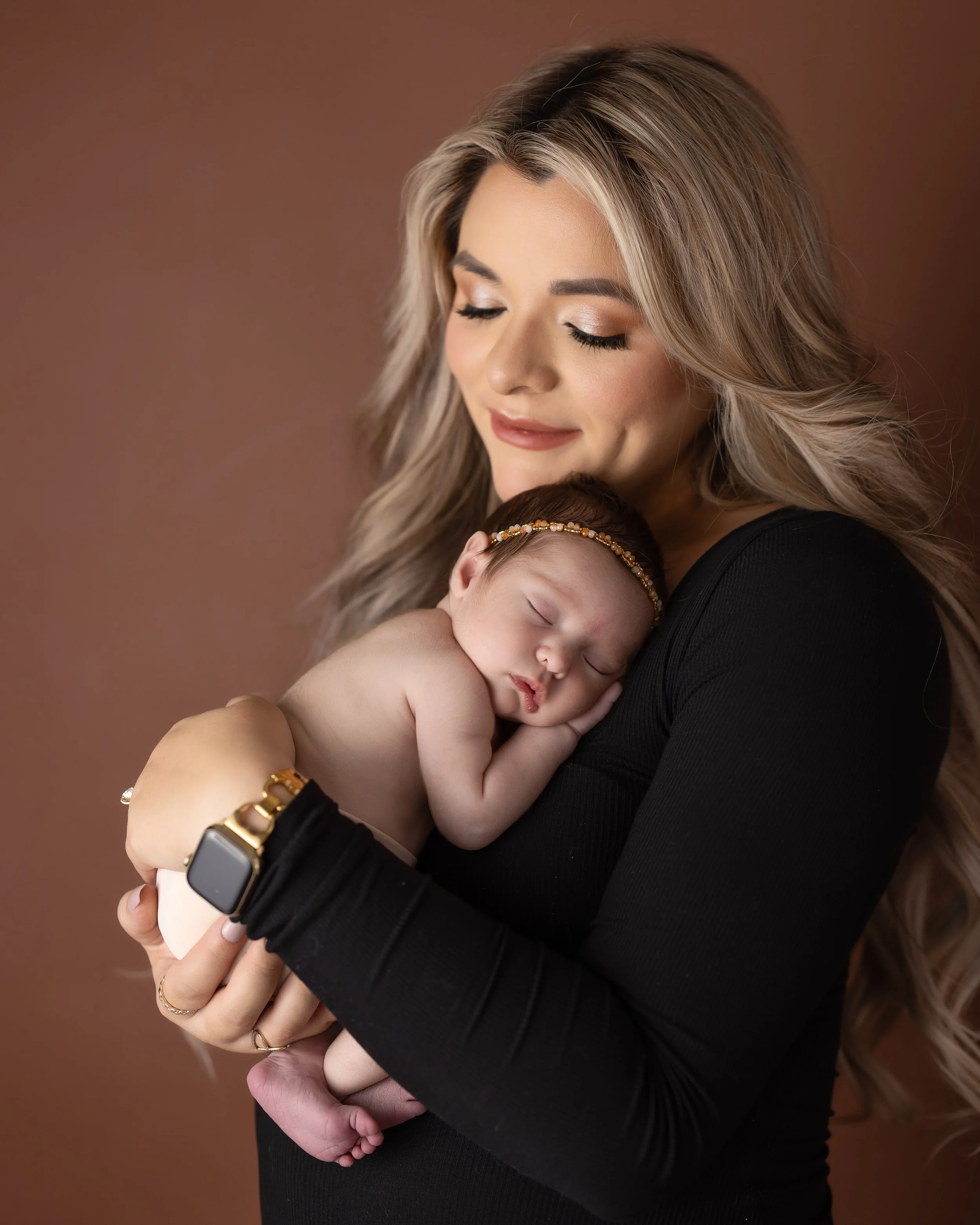 A woman with blonde hair holding a sleeping baby with a gold headband, against a brown background.