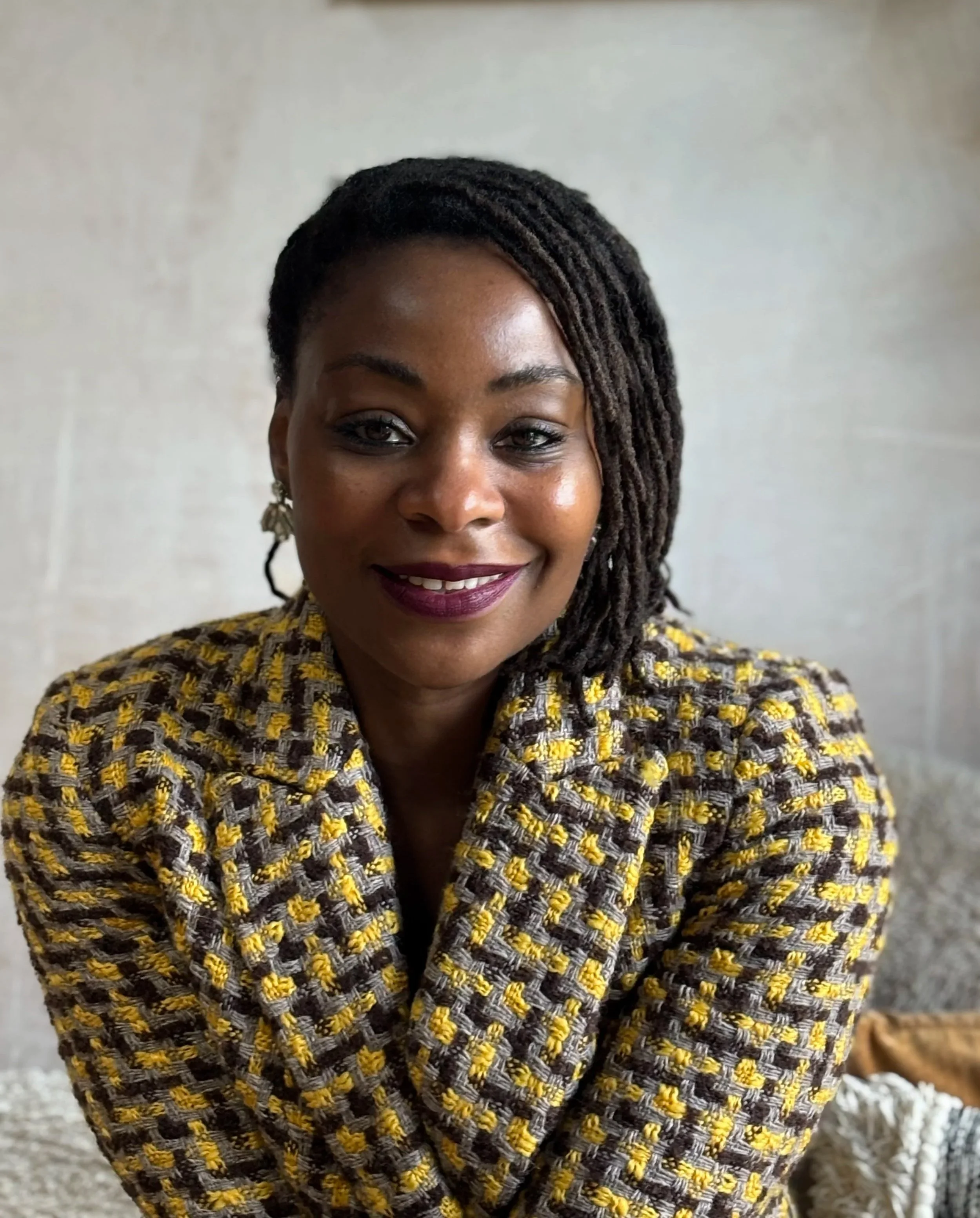 Dr Tosin Bowen-Wright - A black woman with shoulder-length dreadlocks, smiling and wearing a yellow and gray patterned blazer.