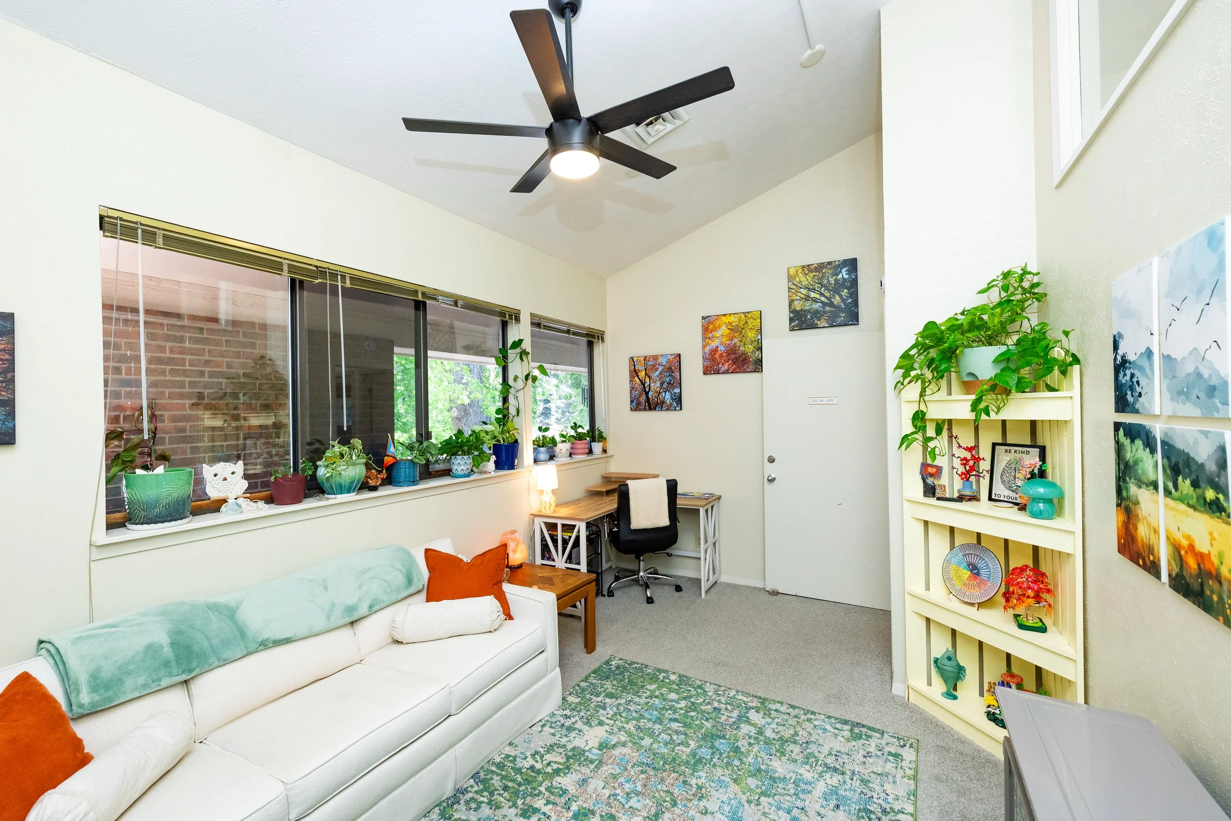 Living room with a white couch, orange pillows, large windows with plants on the windowsill, colorful artwork on the wall, a small desk with a chair, and a yellow bookshelf with decorative items and plants.