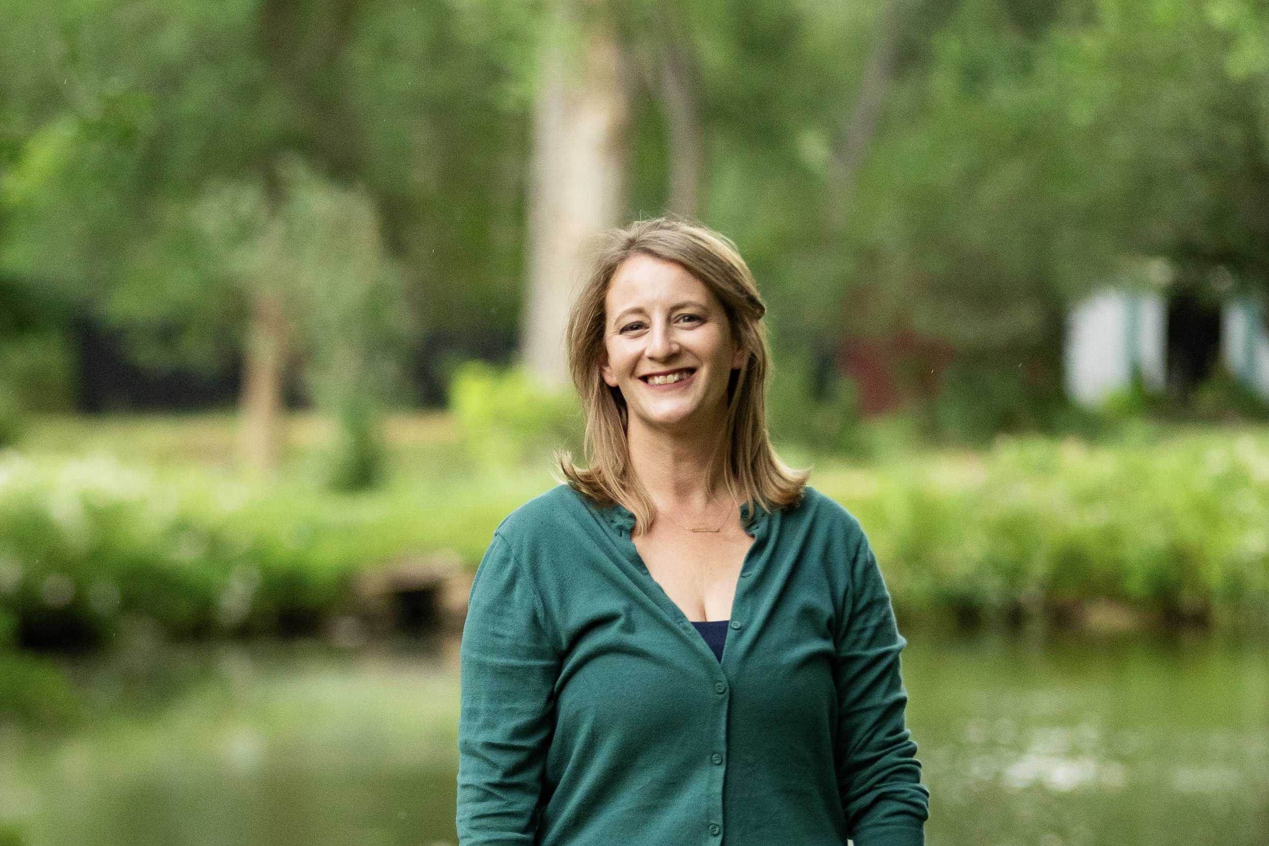 A woman with shoulder-length light brown hair smiling outdoors in a park with green trees in the background, wearing a teal blouse.