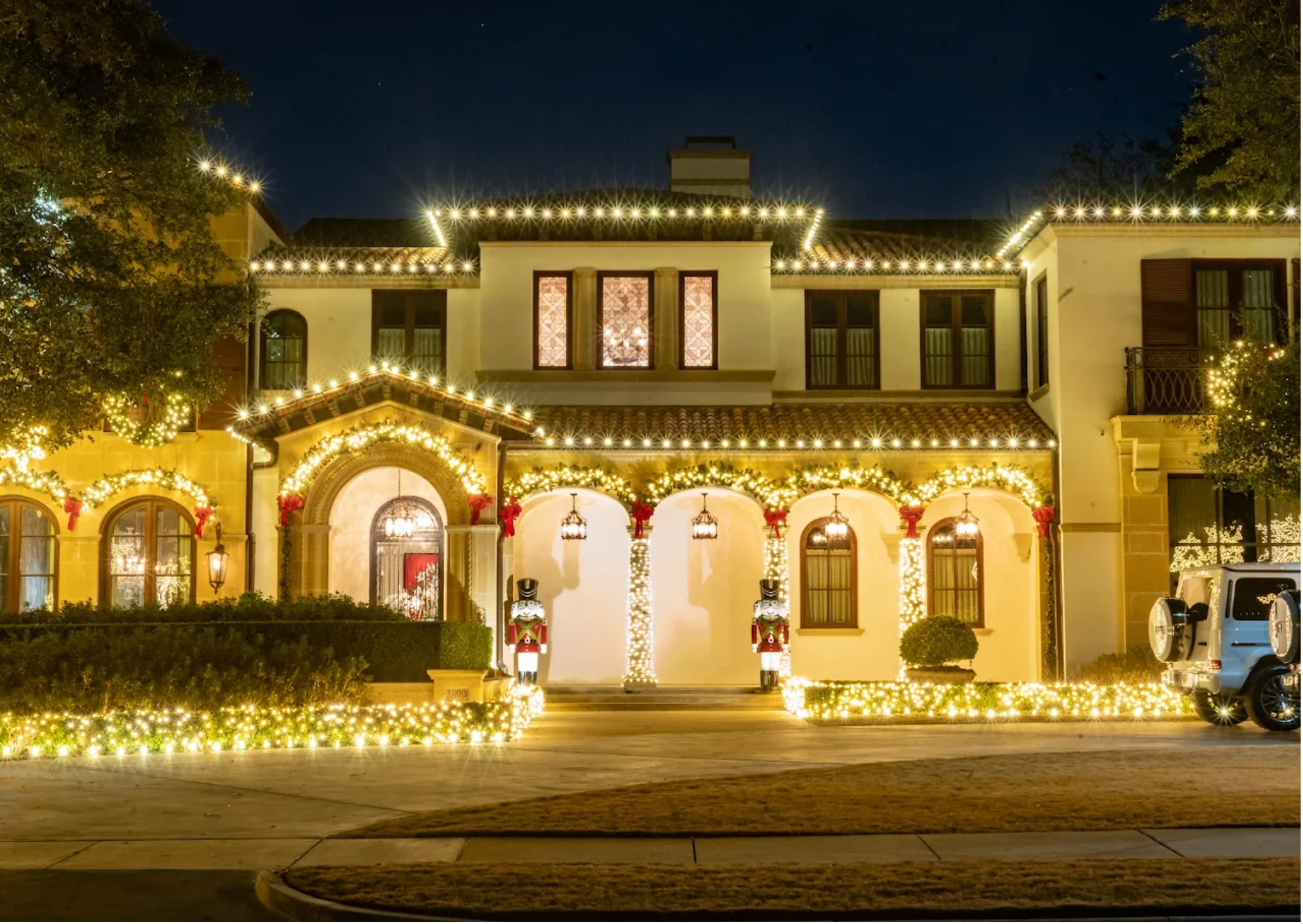 A large, multi-story house decorated with Christmas lights and holiday decorations at night. The house has a Mediterranean style with arched windows and a tiled roof. Nutcracker figures stand at the entrance, and strings of lights outline the roof, windows, and yard. Two tall nutcracker statues are positioned in front of the house, flanking the entrance. The yard features a hedge and a driveway with a parked vehicle.
