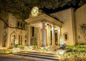 A grand, illuminated entrance of a mansion with tall columns, surrounded by well-kept landscaping and decorative plants at night.