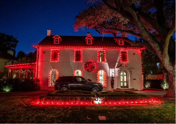 A large white house decorated with red Christmas lights and a wreath on the front door. A black car is parked in the driveway. There is a decorated Christmas tree to the right of the house. A large tree is in the front yard, and the house number 222 is displayed on a small sign in the yard. It is nighttime.