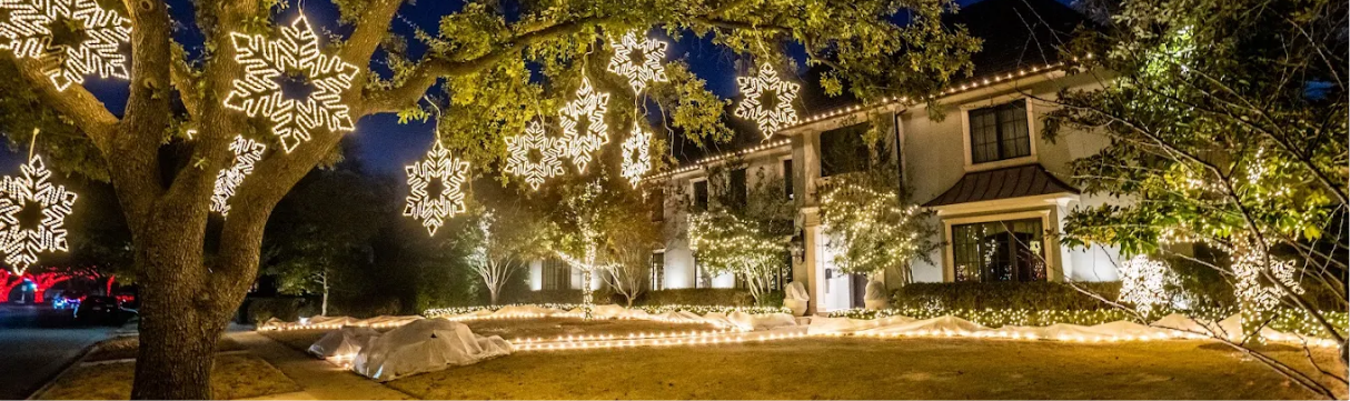 A house decorated with Christmas lights, including snowflake-shaped lights hanging from a large tree and lights outlining the house and yard at night.