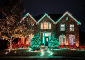 A large brick house decorated with colorful Christmas lights at night, with lit-up bushes and pathway lights leading to the front door.