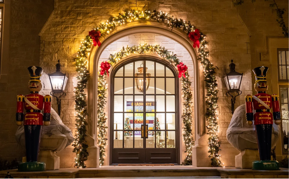 Festively decorated house entrance with an arched doorway, surrounded by Christmas garland, lights, and red poinsettias, flanked by two nutcracker statues.