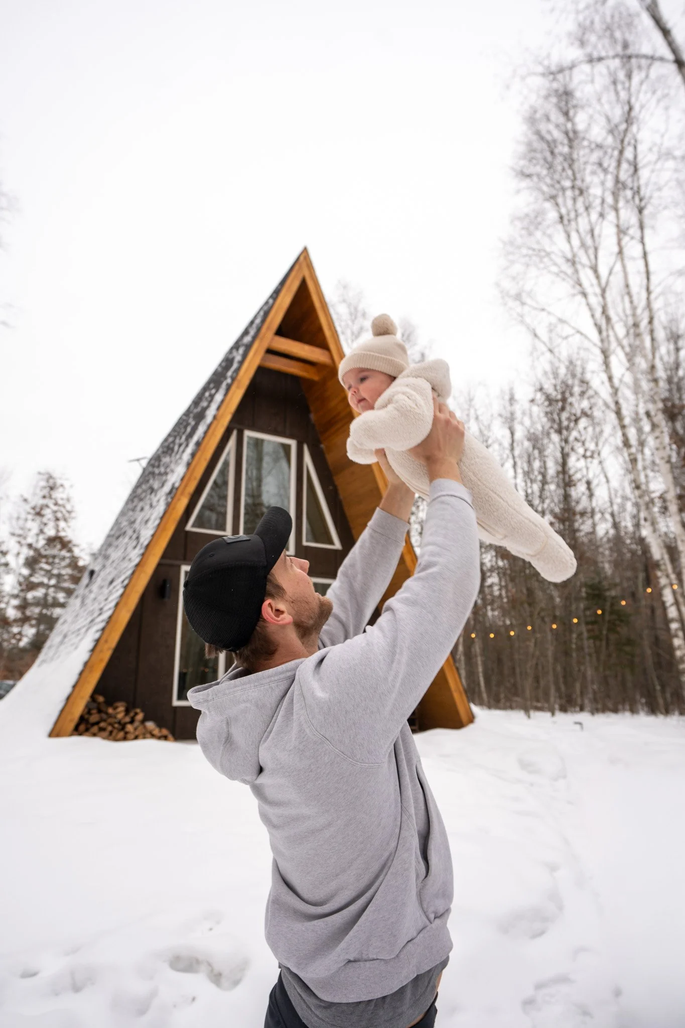 A man lifting a baby in the snow outdoors in front of a triangular A-frame cabin with large windows, surrounded by leafless trees.
