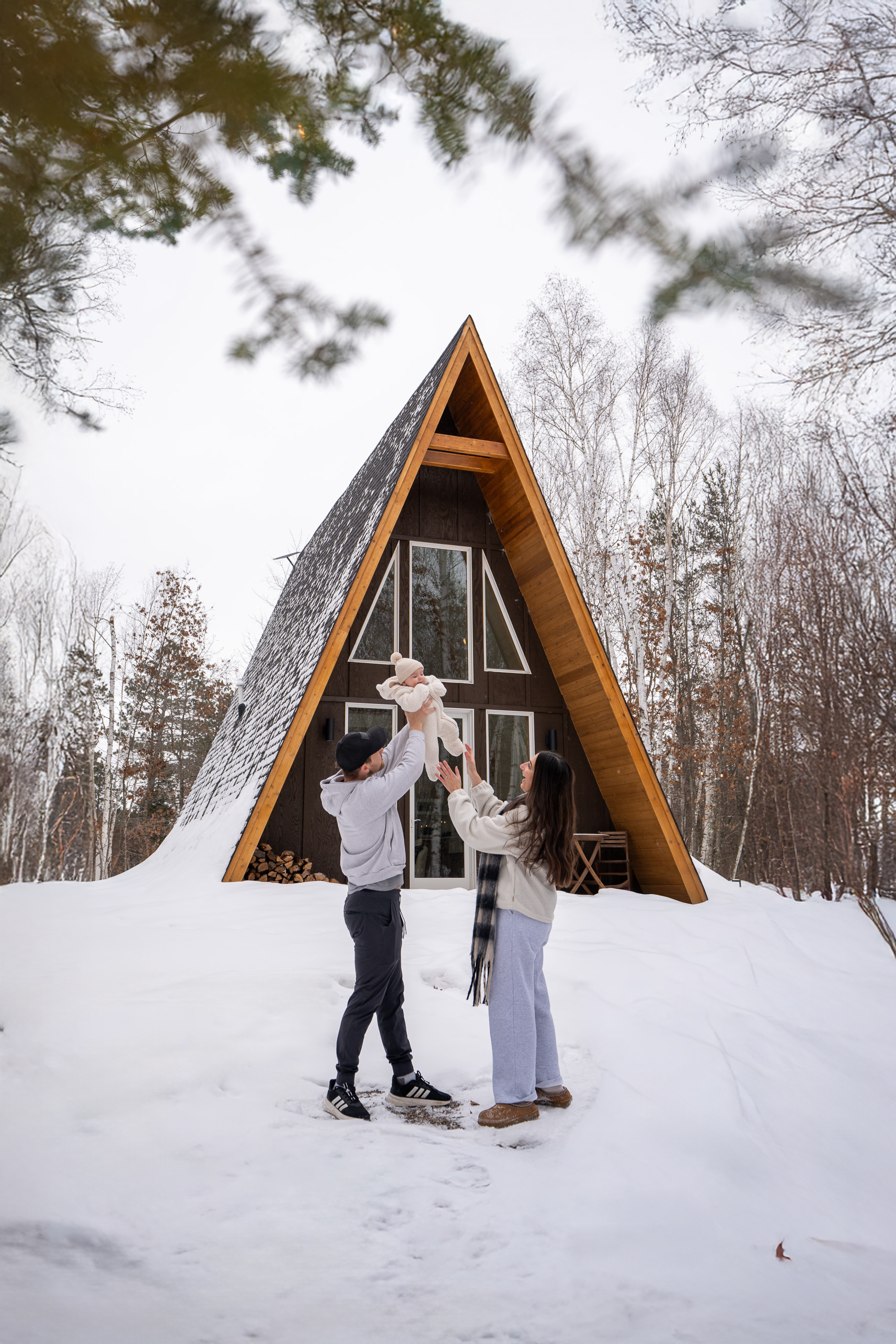 A family of three playing in the snow in front of a modern A-frame house in winter, with bare trees in the background.