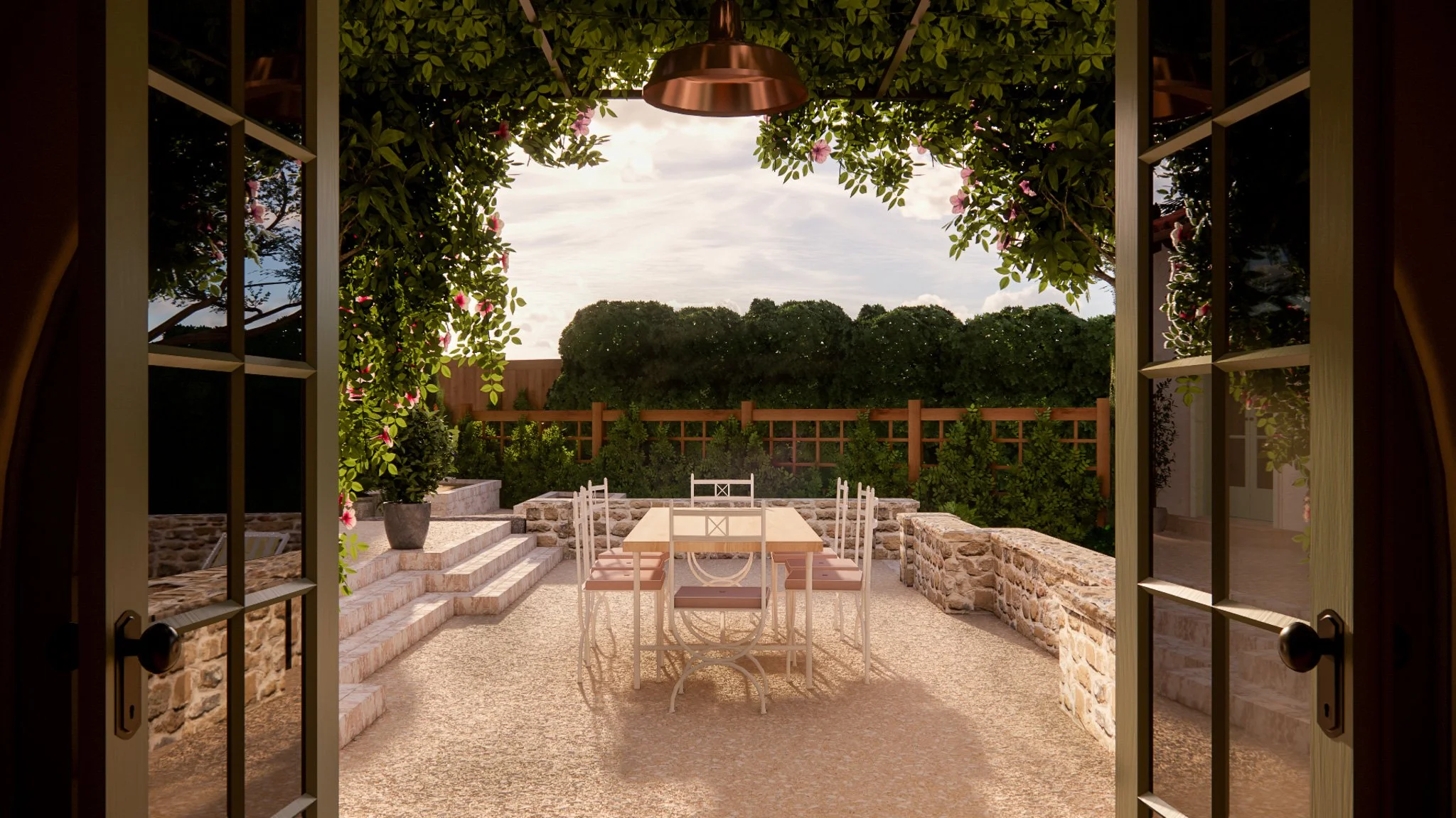 View of a cozy outdoor patio with a wooden dining table and chairs, surrounded by stone and brick walls, framed by open French doors, lush green bushes, and flowering plants, under a partly cloudy sky.
