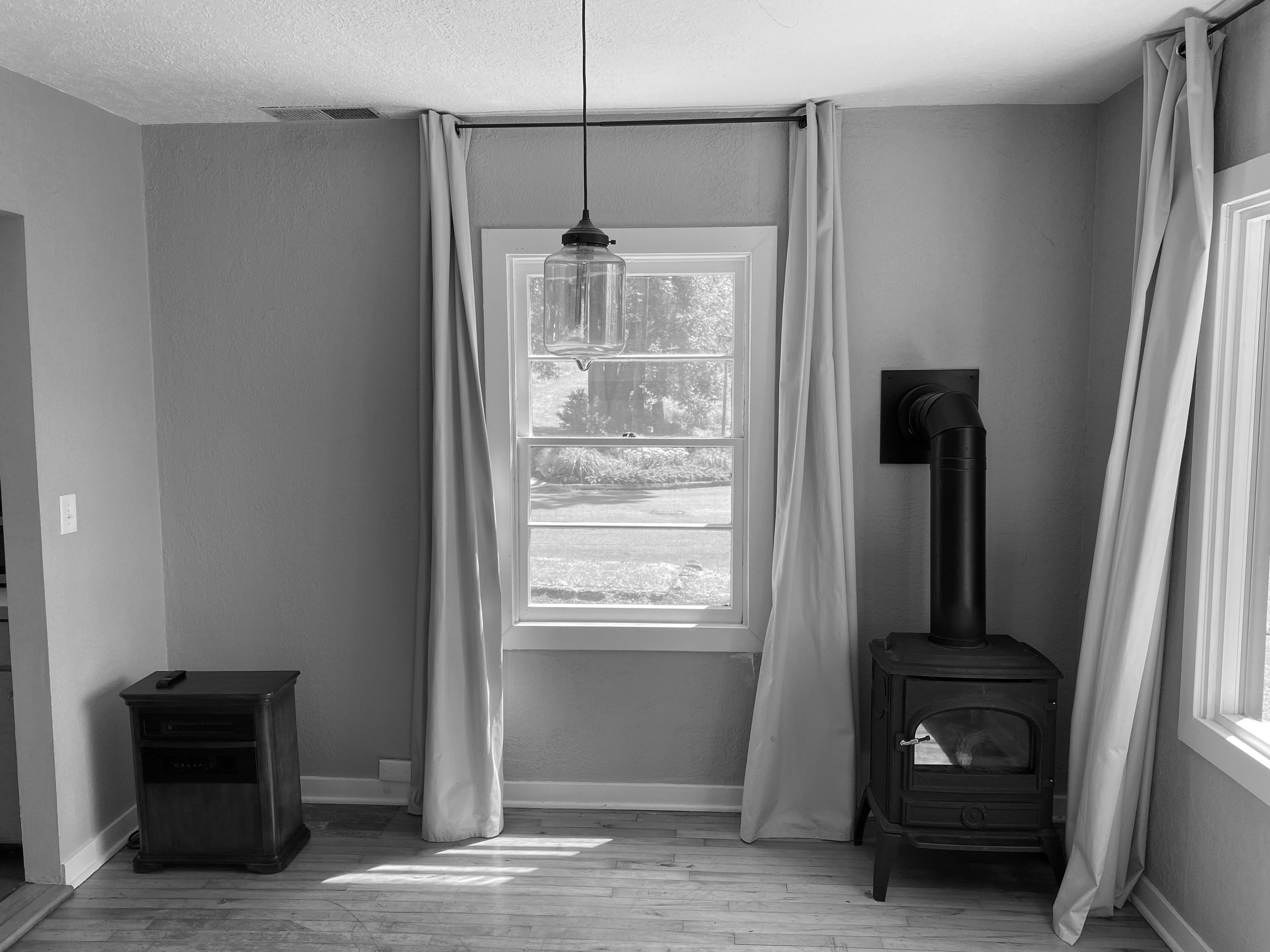 Black and white photo of a room with a wood-burning stove, window with curtains, and a small wooden cabinet on a hardwood floor.