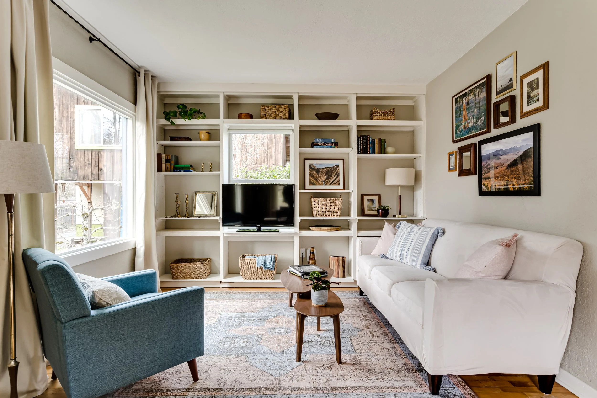 Cozy living room with white couch, blue armchair, wooden coffee table, wall-mounted TV, bookshelves with decor, and large window.