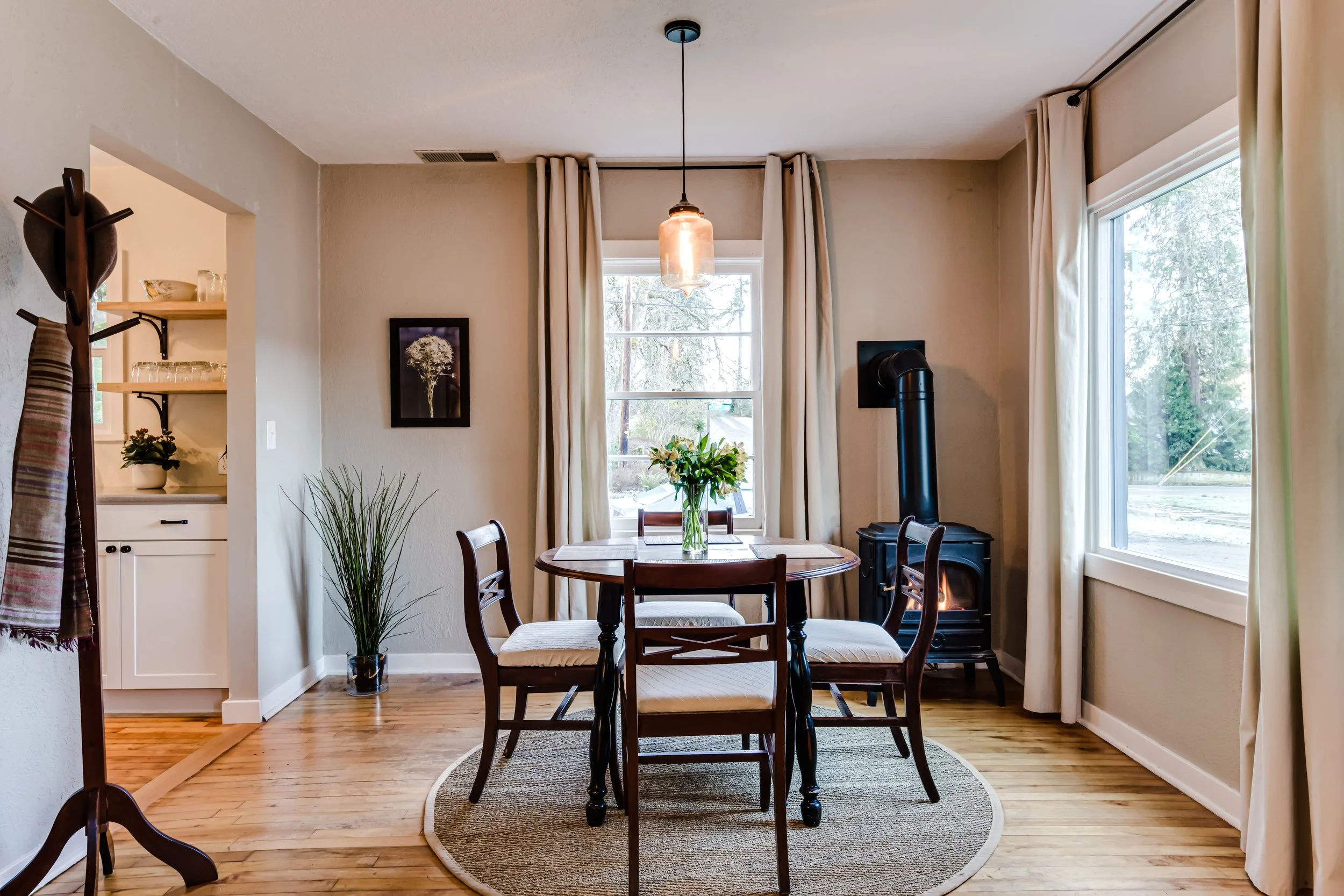 Cozy dining room with round table, four chairs, fireplace, large windows, hanging light, and framed wall art.