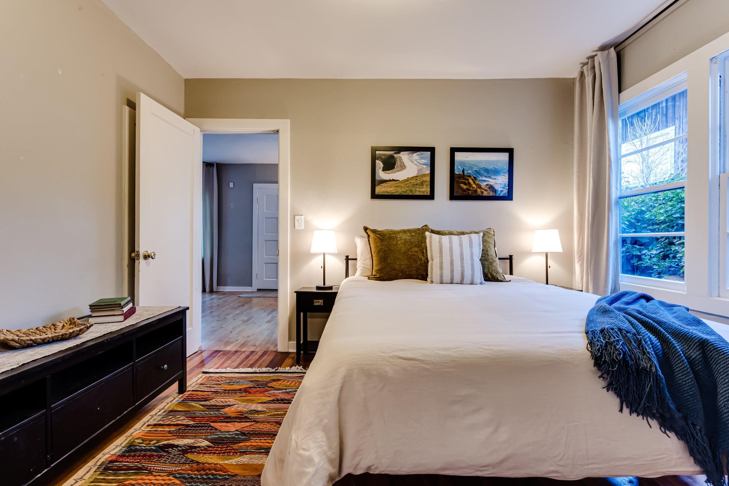 Cozy bedroom with a neatly made bed, decorative pillows, and a blue throw. Two landscape pictures hang above the bed. Nightstands with lamps on either side. A colorful patterned rug on wooden floor. Window with curtain and outside greenery visible.